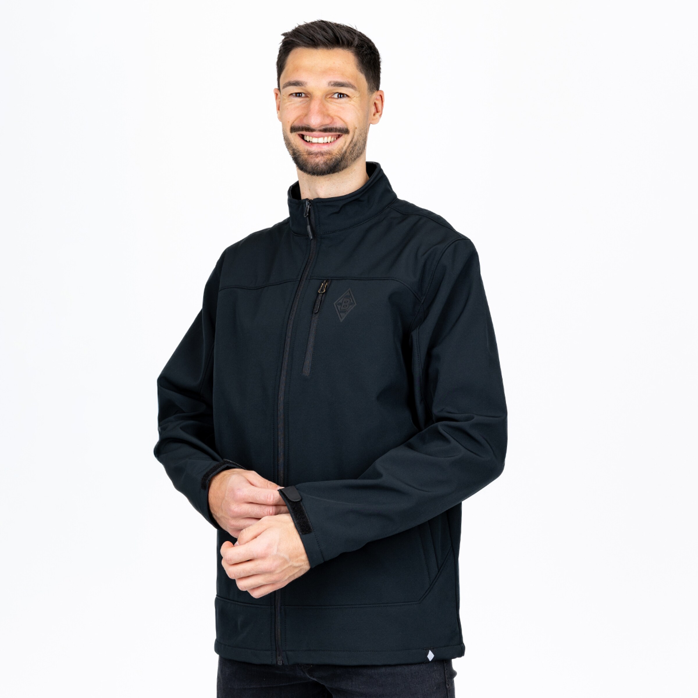 Lächelnder Mann mit Bart in schwarzer Softshelljacke vor weißem Hintergrund. Er justiert den Klettverschluss am Ärmel.