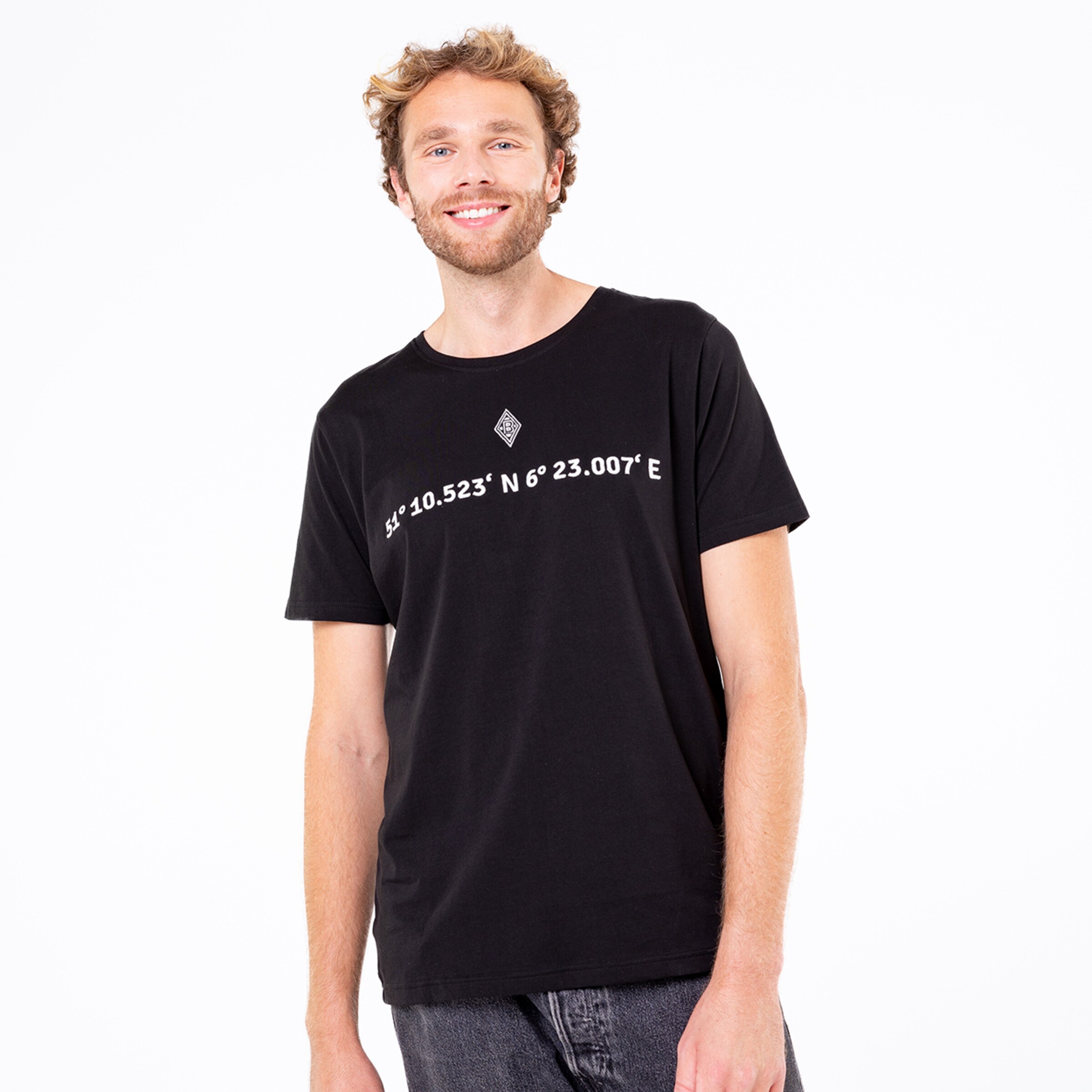 Mann mit lockigen Haaren trägt ein schwarzes T-Shirt mit Koordinaten.