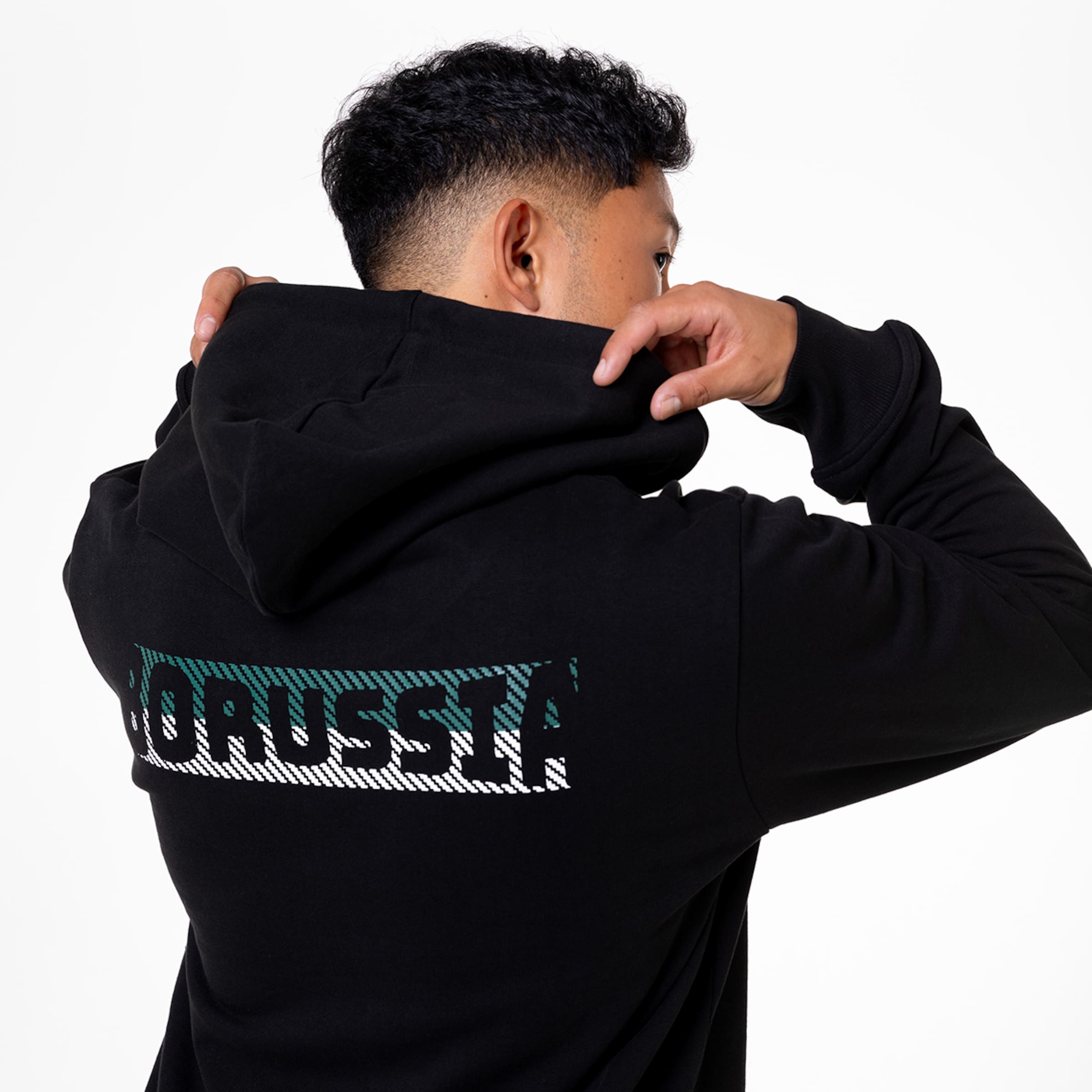 Rückansicht eines Mannes im schwarzen Hoodie mit grün-weißem „BORUSSIA“-Schriftzug. Er zieht die Kapuze hoch. Vor weißem Hintergrund.