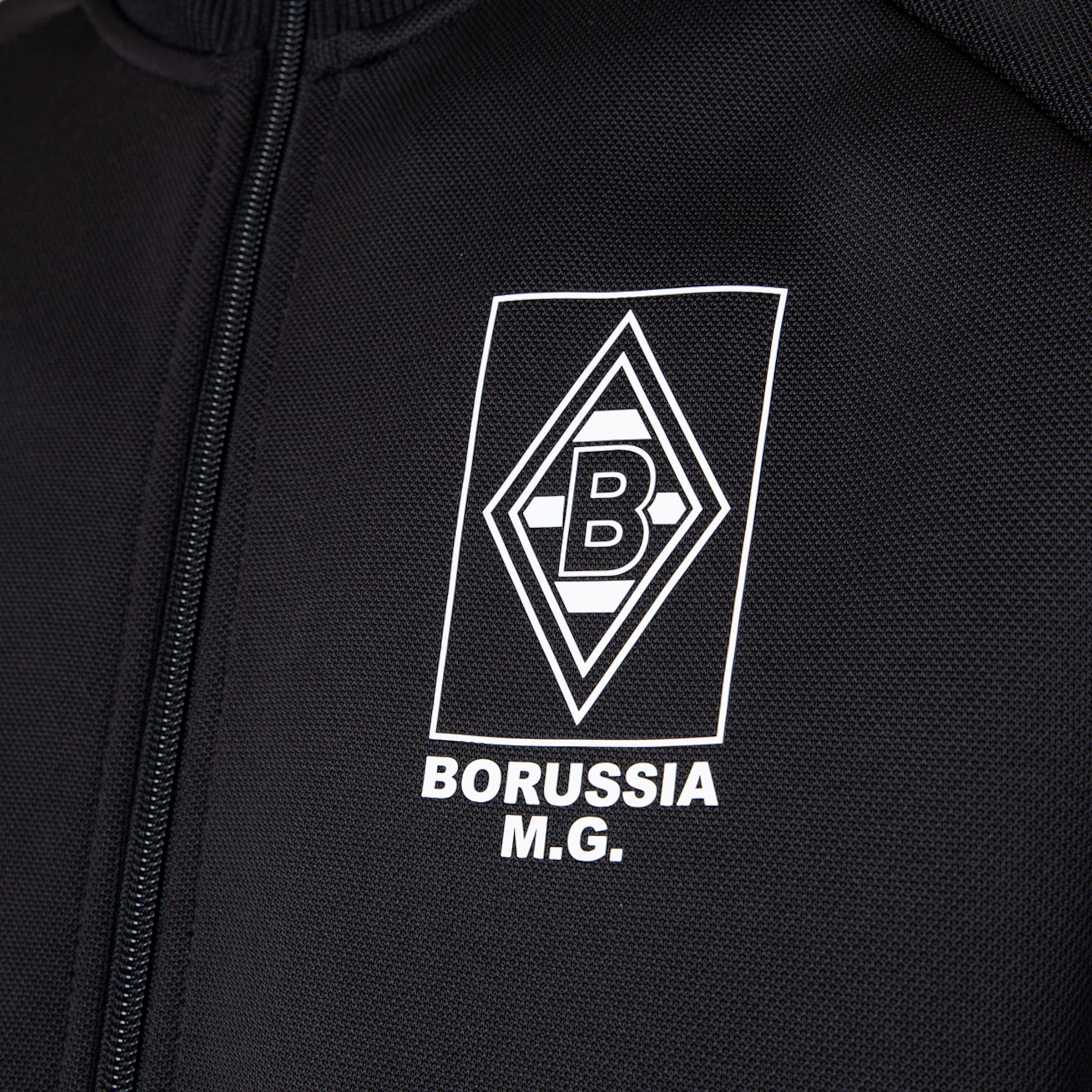 Nahaufnahme einer schwarzen Trainingsjacke mit dem weißen Logo von Borussia Mönchengladbach und dem Schriftzug „BORUSSIA M.G.“