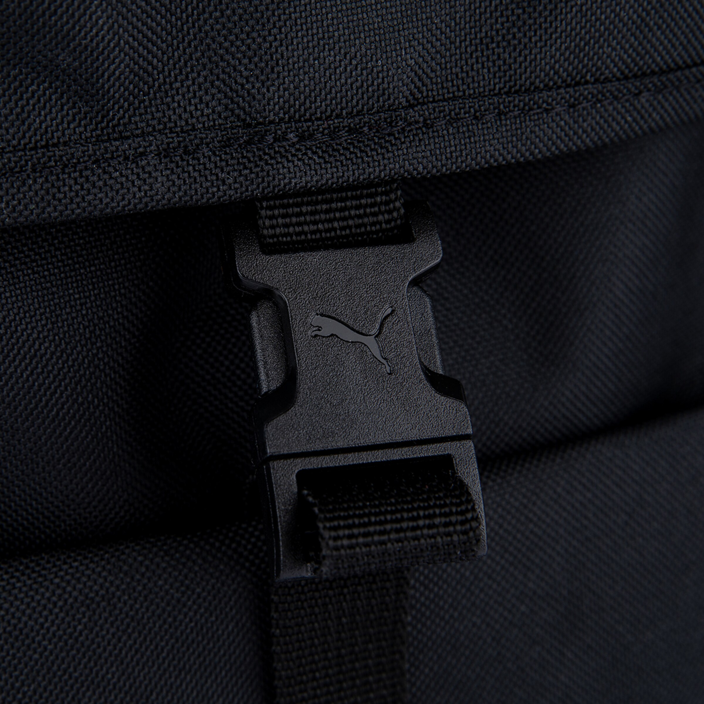 Nahaufnahme eines schwarzen strukturierten Stoffes, vermutlich von einem Rucksack oder einer Tasche. Im Zentrum ist eine schwarze Kunststoffschnalle mit dem eingeprägten Puma-Logo zu sehen. Der Stoff weist eine deutlich sichtbare Webstruktur auf.