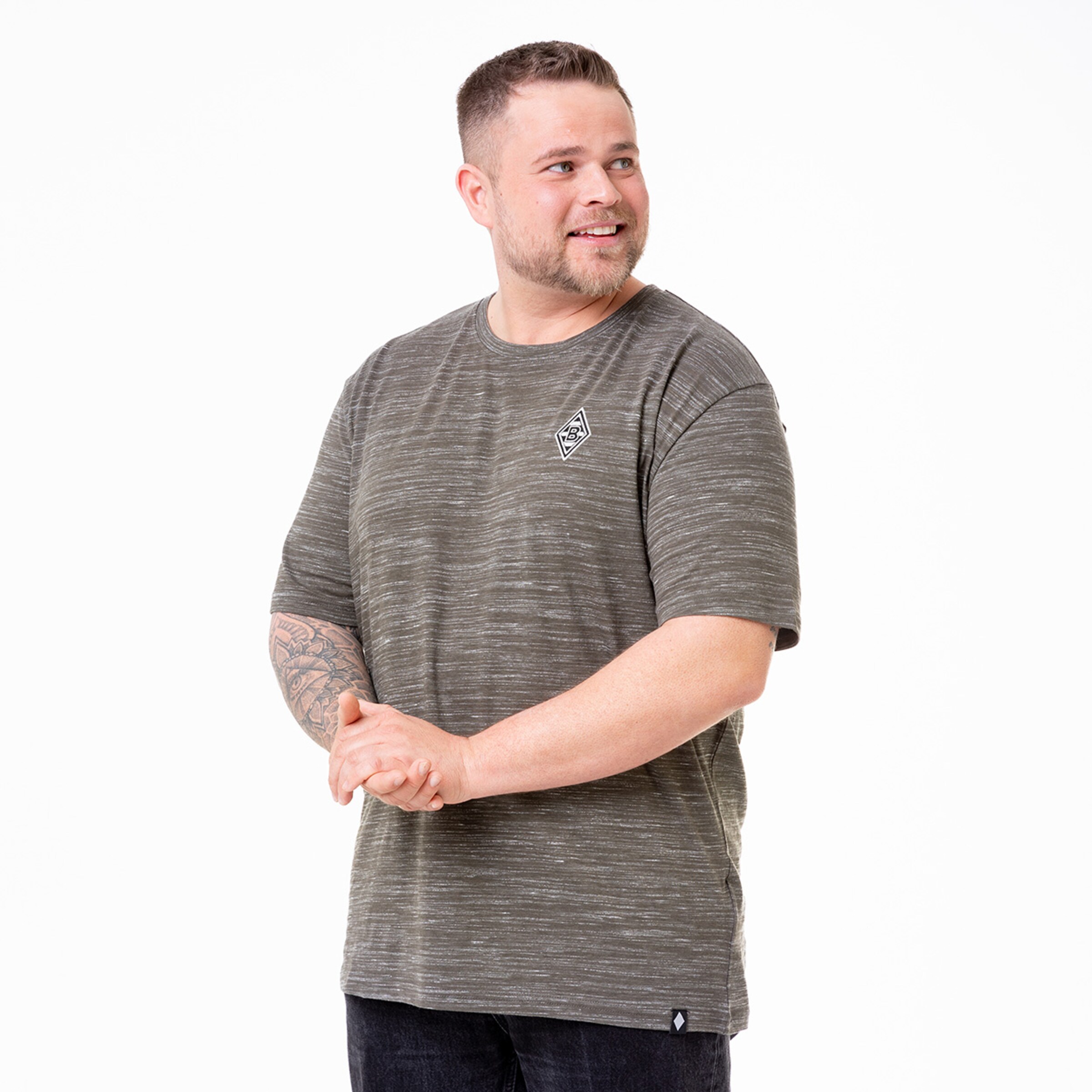 Mann mit tätowierten Armen steht lächelnd in einem grauen gestreiften T-Shirt.