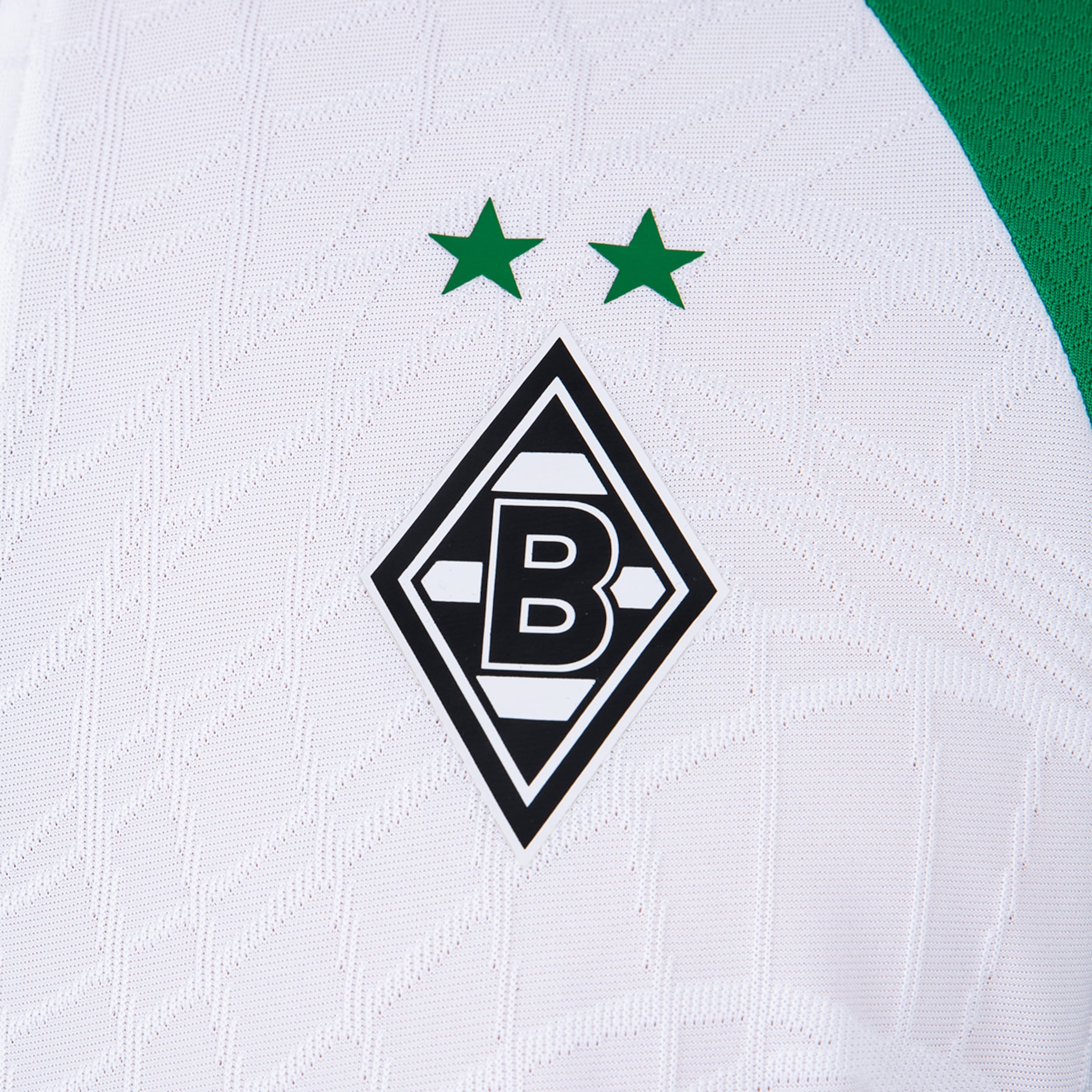 Nahaufnahme eines weißen Fußballtrikots von Borussia Mönchengladbach mit dem schwarzen Rautenlogo, einem weißen 'B' und zwei grünen Sternen. Das Trikot zeigt dezente Texturen und einen grünen Bereich auf der rechten Seite.