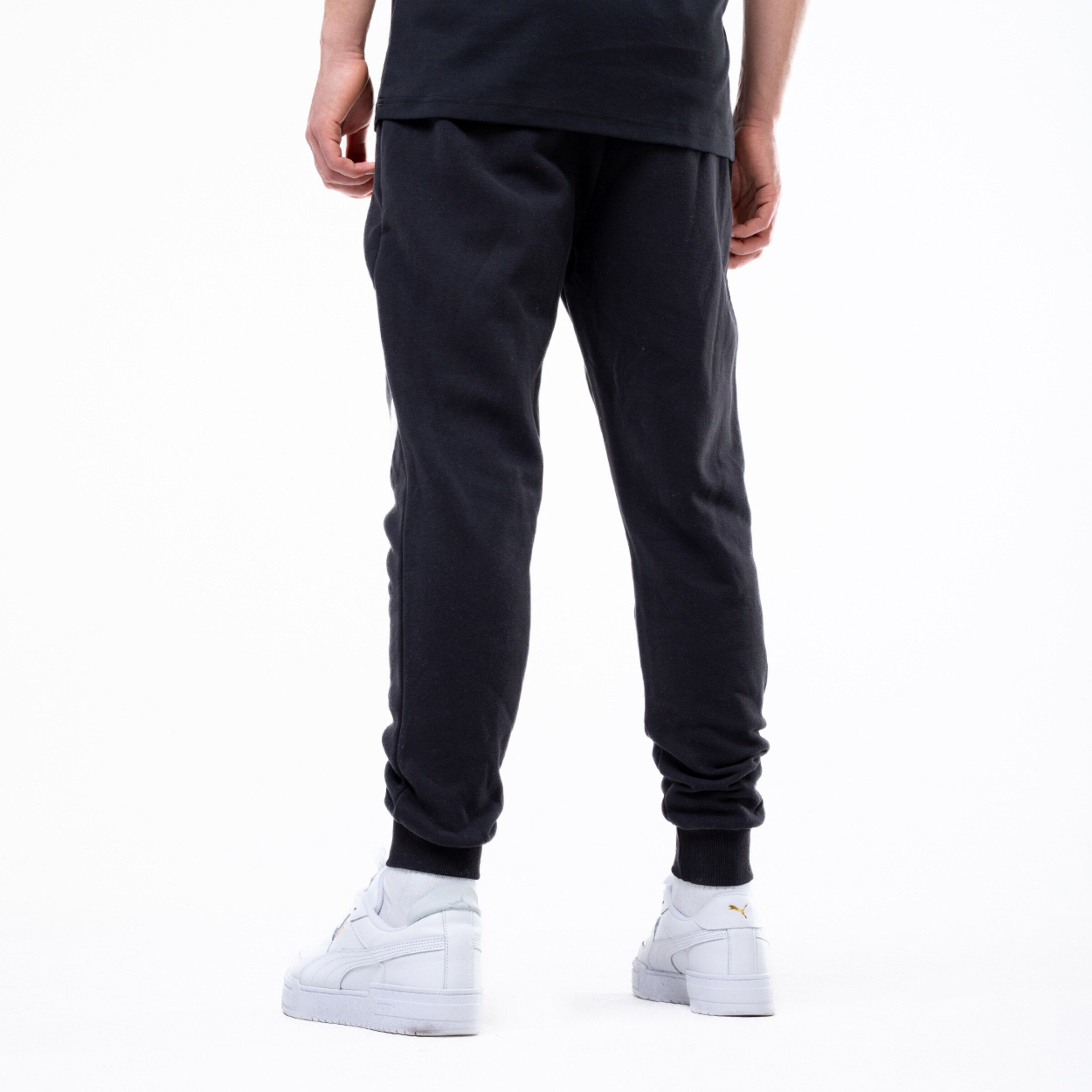 Rückansicht einer Person in schwarzer Jogginghose, schwarzem T-Shirt und weißen Puma-Sneakern vor weißem Hintergrund.