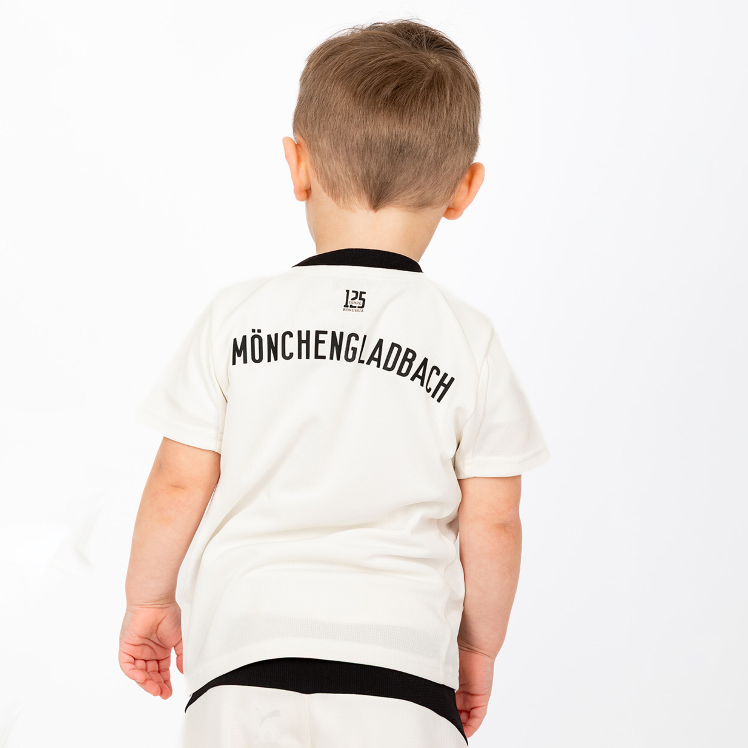 Ein kleines Kind das von hinten in einem weißen Borussia Mönchengladbach T-Shirt mit der Aufschrift MÖNCHENGLADBACH auf dem Rücken zu sehen ist.