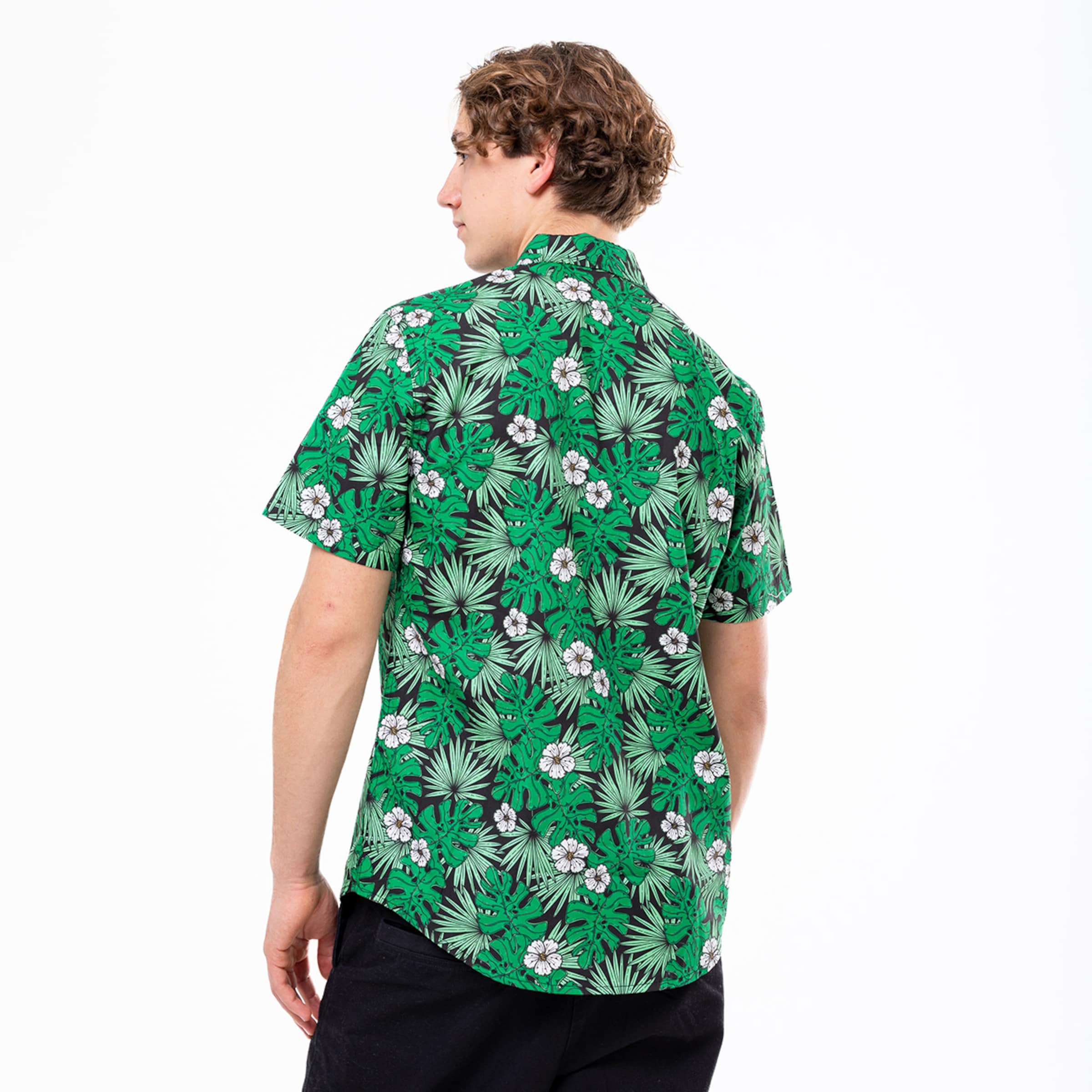 Ein Mann von hinten in einem grünen Kurzarmhemd mit tropischem Muster und schwarzer Hose vor weißem Hintergrund.