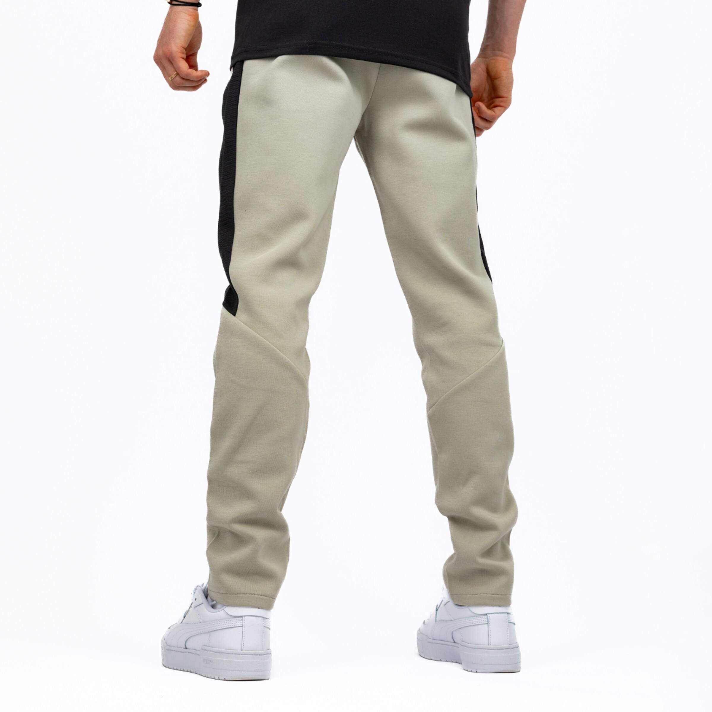 Eine Person, von der Taille abwärts und von hinten gesehen, trägt hellbeige Jogginghosen mit schwarzen Streifen an den Seiten und weiße Sneaker, vor einem reinweißen Hintergrund stehend.