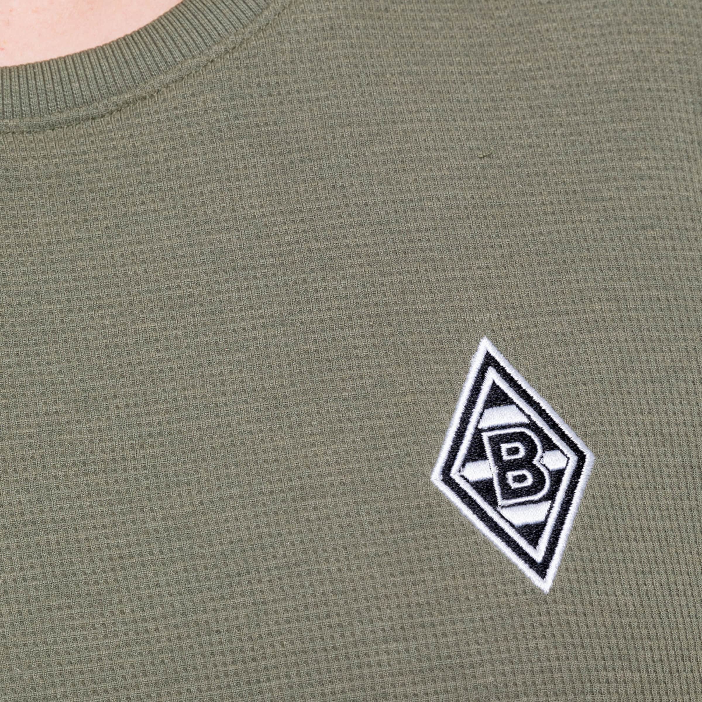 Nahaufnahme eines olivgrünen Oberteils mit gesticktem Borussia Mönchengladbach-Logo.