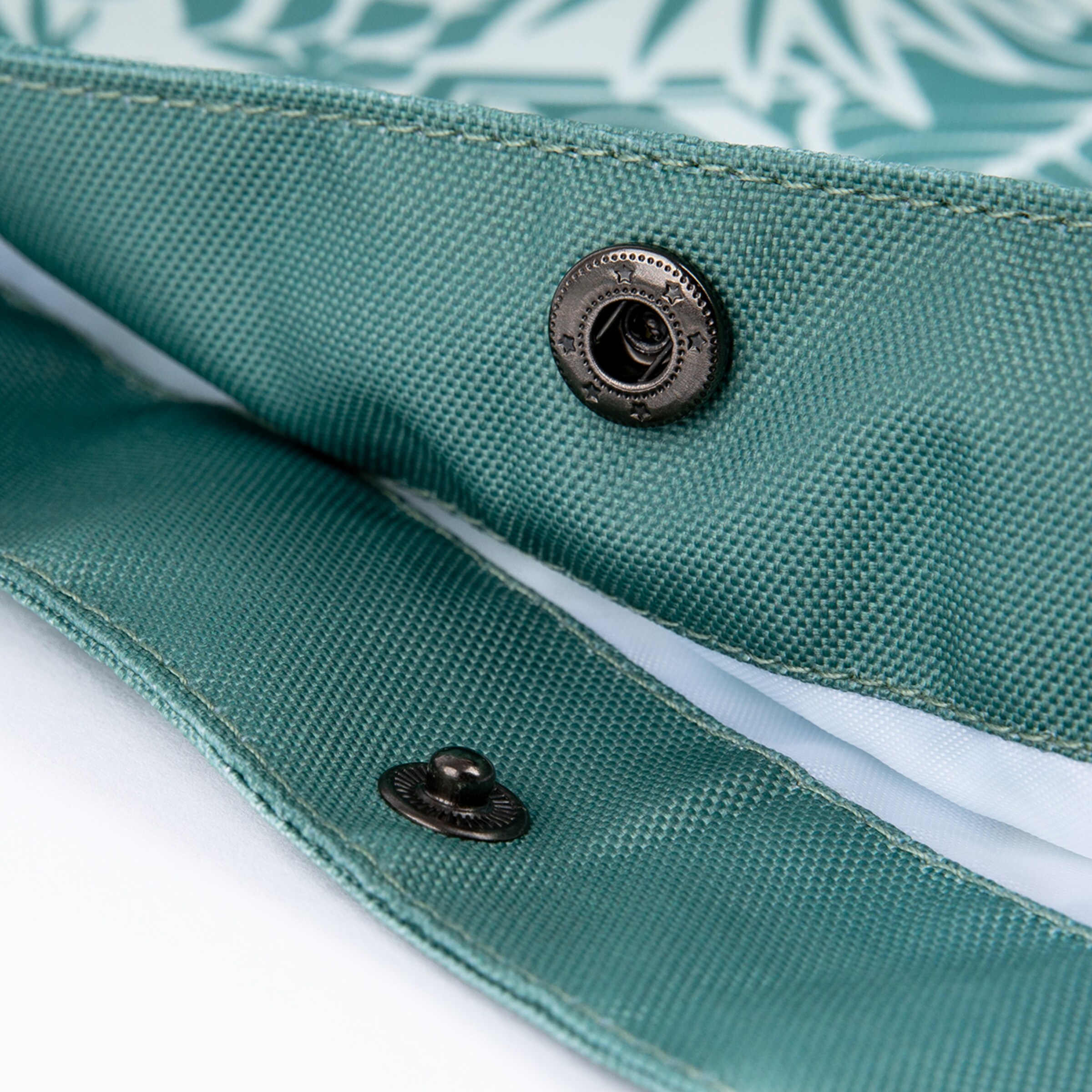 Nahaufnahme eines türkisfarbenen Gewebes, möglicherweise einer Tasche, mit zwei dunklen metallischen Druckknöpfen. Ein Druckknopf hat ein Sternenmuster. Helle Nähte sind sichtbar, vor unscharfem hellem Hintergrund.