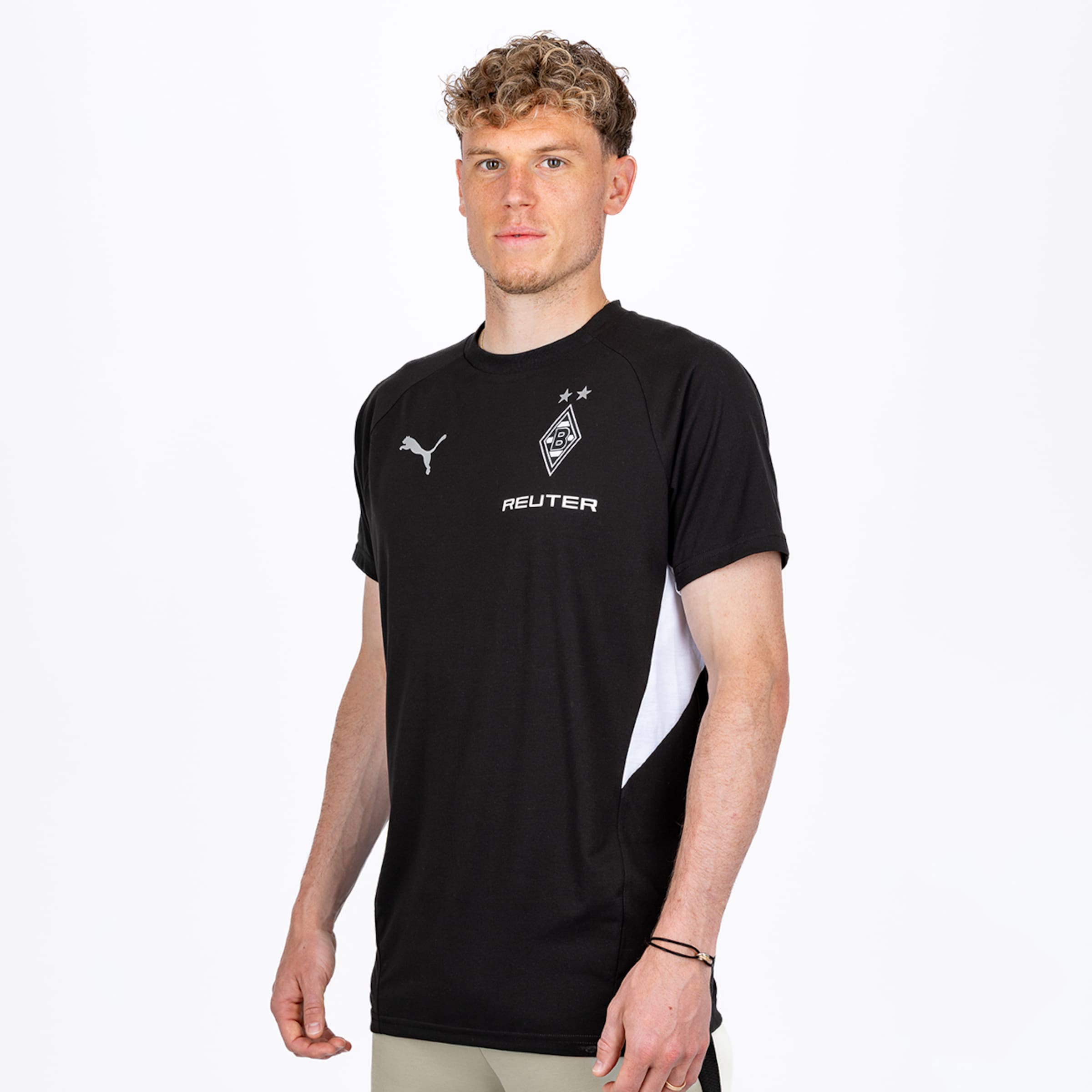 Ein Mann mit lockigem blondem Haar posiert in einem schwarzen Sport-T-Shirt mit Puma, Borussia Mönchengladbach-Wappen, zwei Sternen und dem Sponsor „REUTER“ auf weißem Hintergrund.
