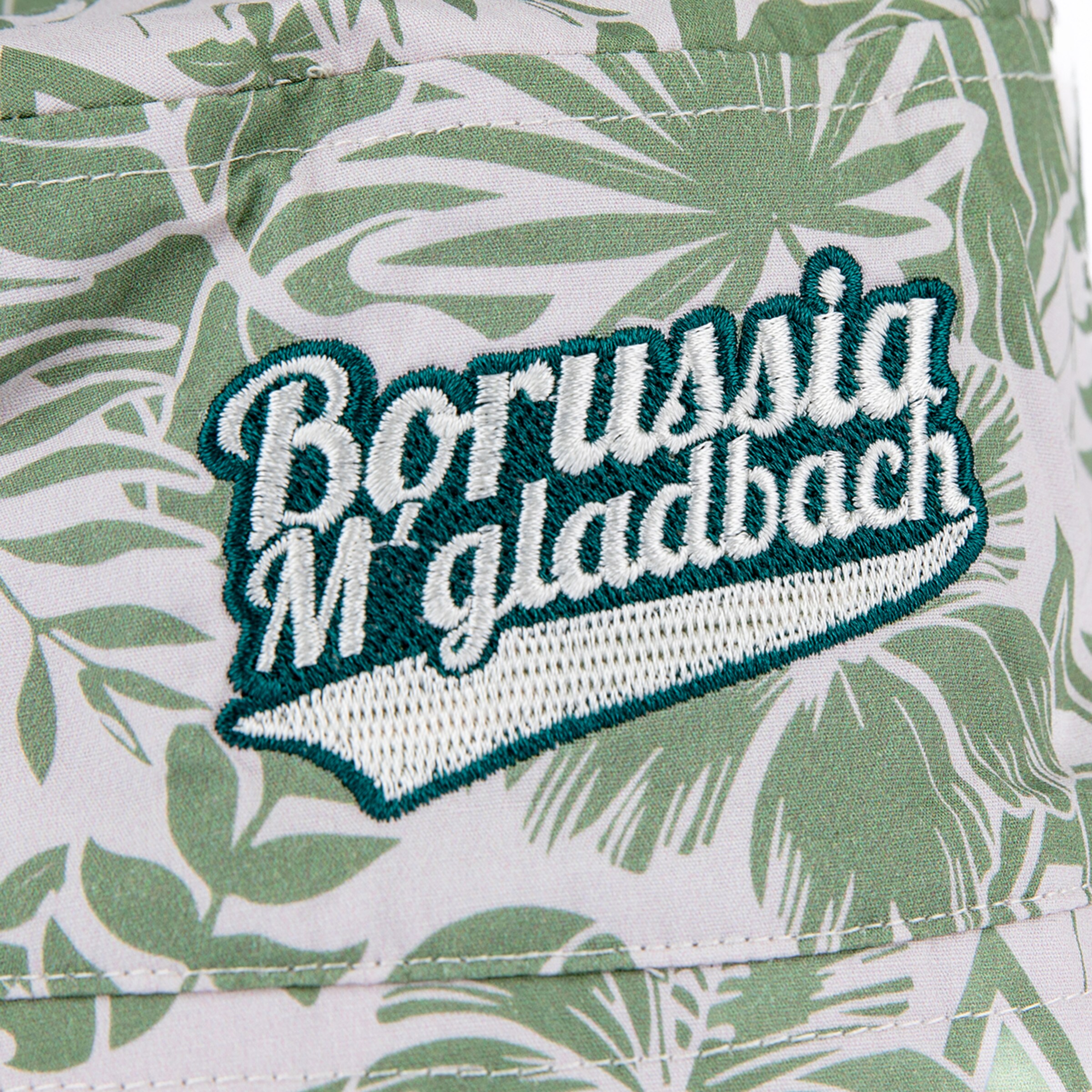 Nahaufnahme eines hellen Stoffes mit grünem tropischen Blattmuster, auf dem ein gesticktes 'Borussia M'gladbach'-Logo zu sehen ist. Der Schriftzug ist weiß und mit dunkelgrüner Stickerei umrandet und hinterlegt, was sich vom gemusterten Stoff abhebt.