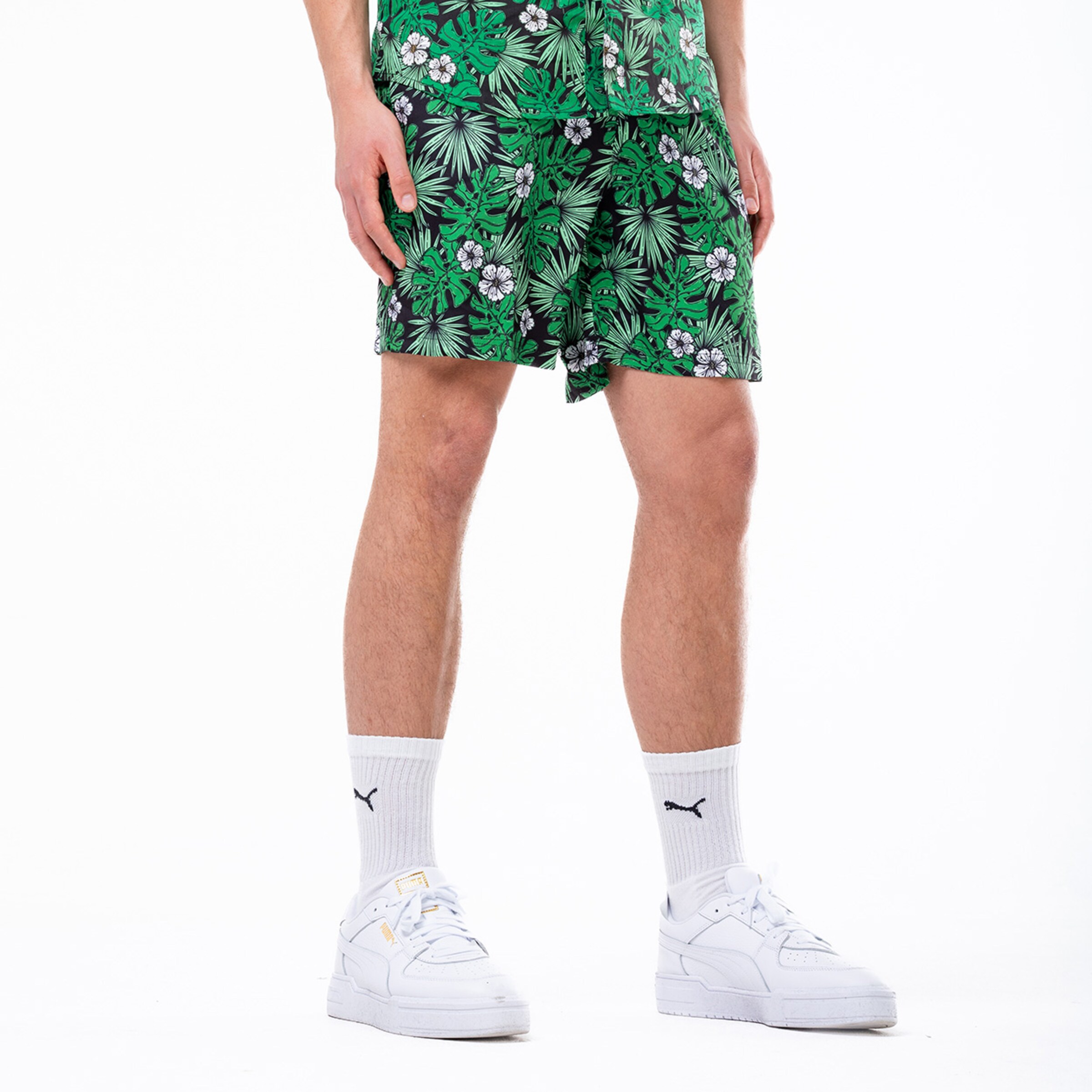 Mann steht in grüner Blumenschorts mit passendem Oberteil und weißen Sneakers.