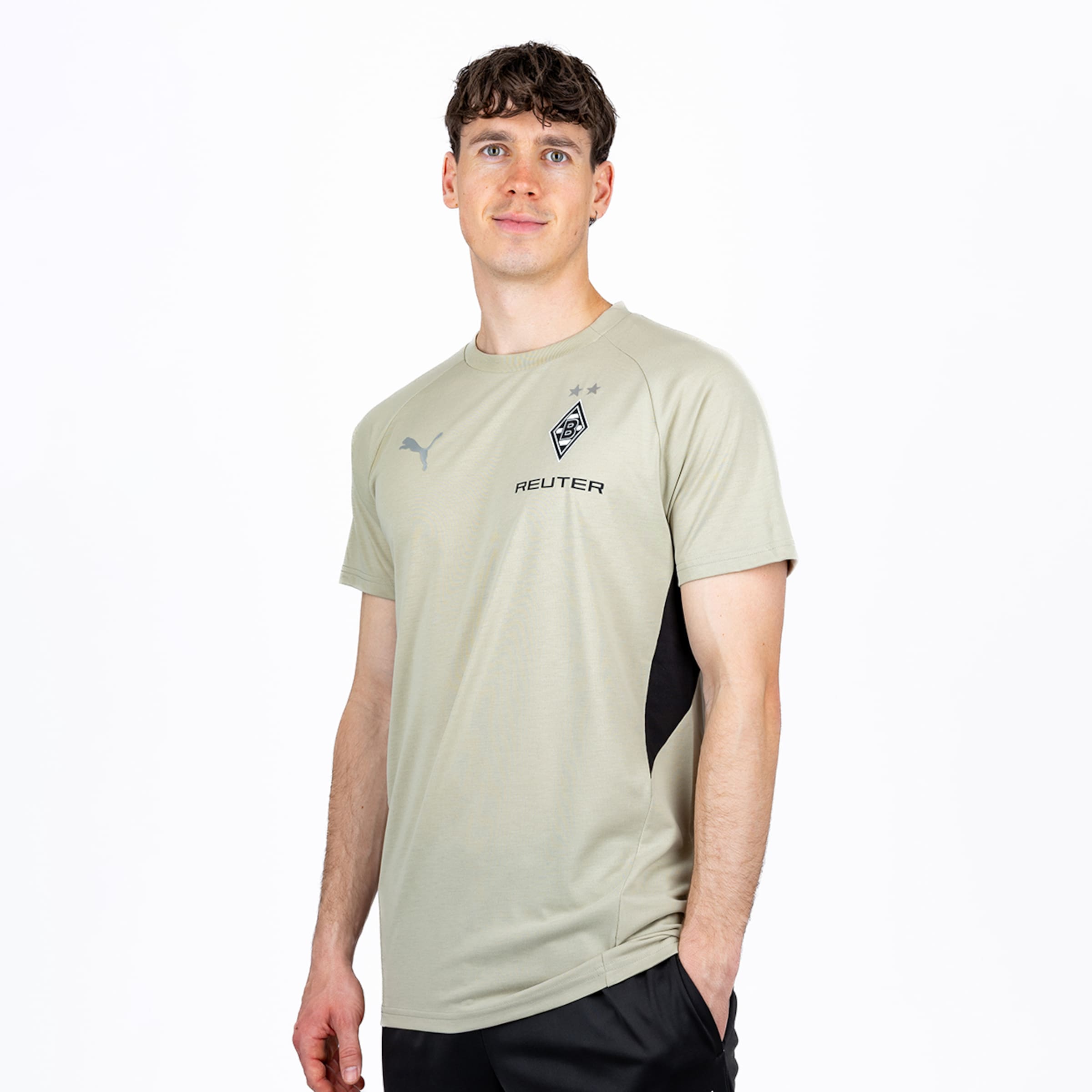 Ein junger Mann mit lockigem Haar steht lächelnd vor weißem Hintergrund. Er trägt ein hellgrünes Kurzarm-T-Shirt mit Puma-, Borussia Mönchengladbach- und REUTER-Logos sowie schwarze Shorts.