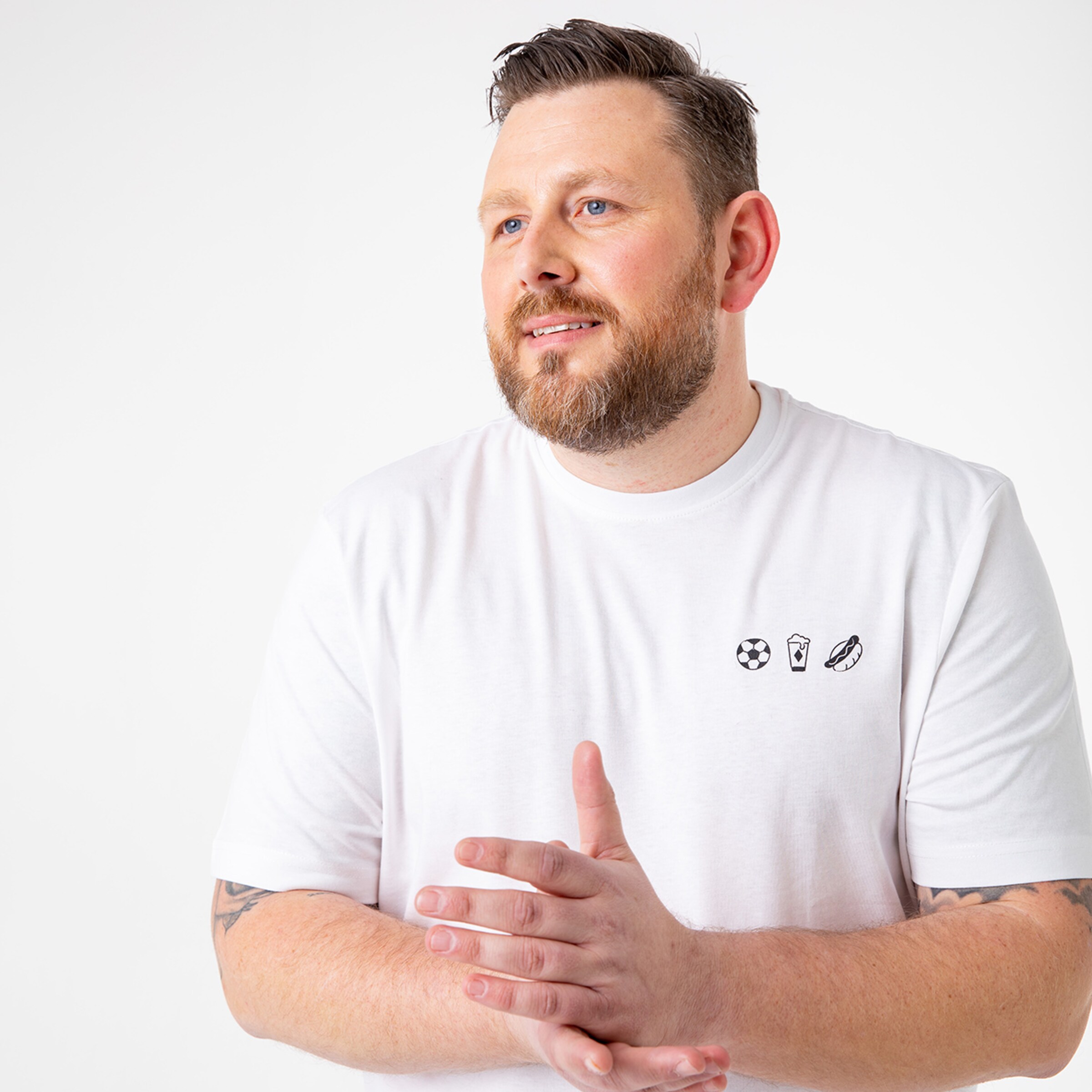 Mann mit Bart trägt ein weißes T-Shirt mit grafischen Motiven. Er steht in nachdenklicher Pose.