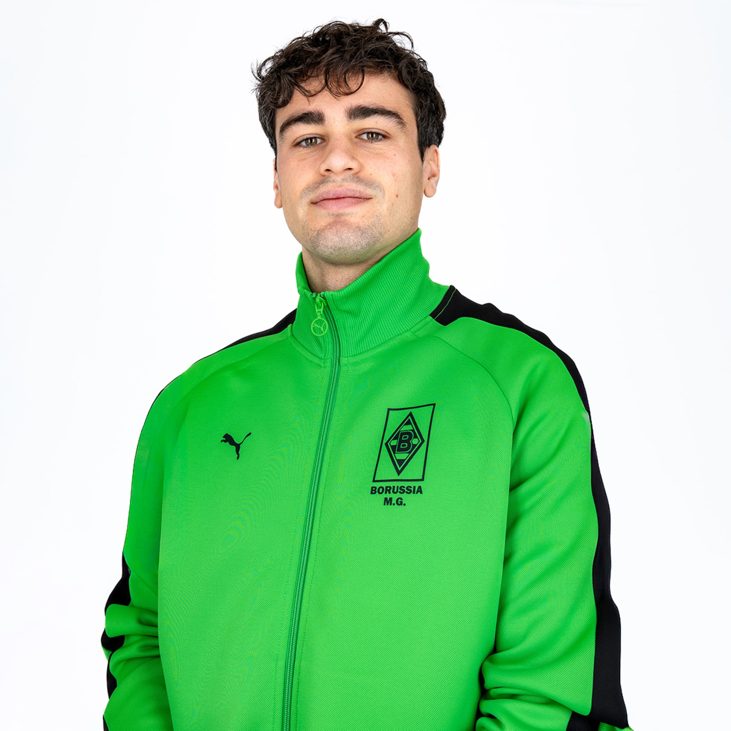Porträt eines Mannes mit dunklen Locken in einer grünen Puma-Trainingsjacke von Borussia Mönchengladbach vor weißem Hintergrund.