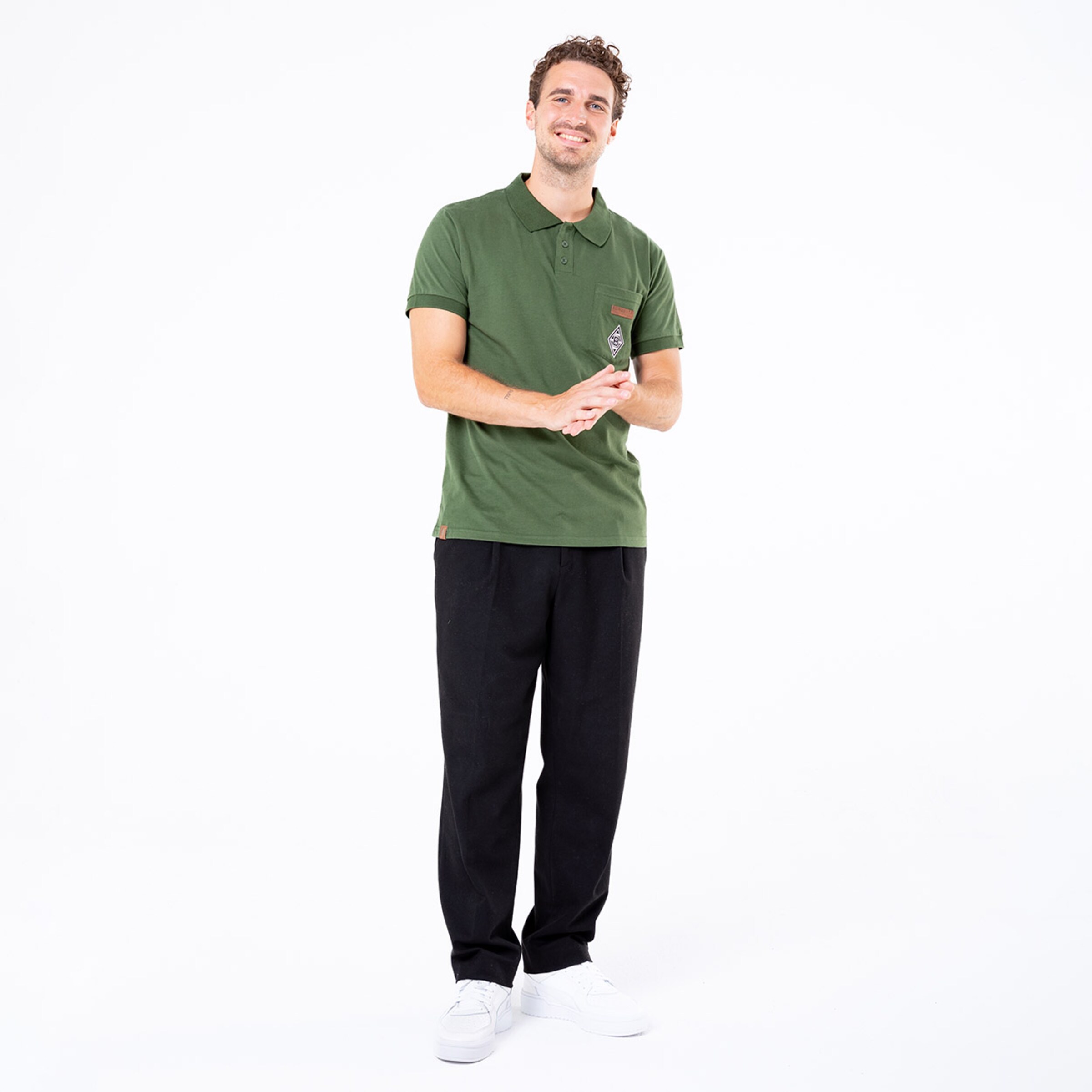 Man wearing a green polo shirt and black pants smiling while holding a phone.