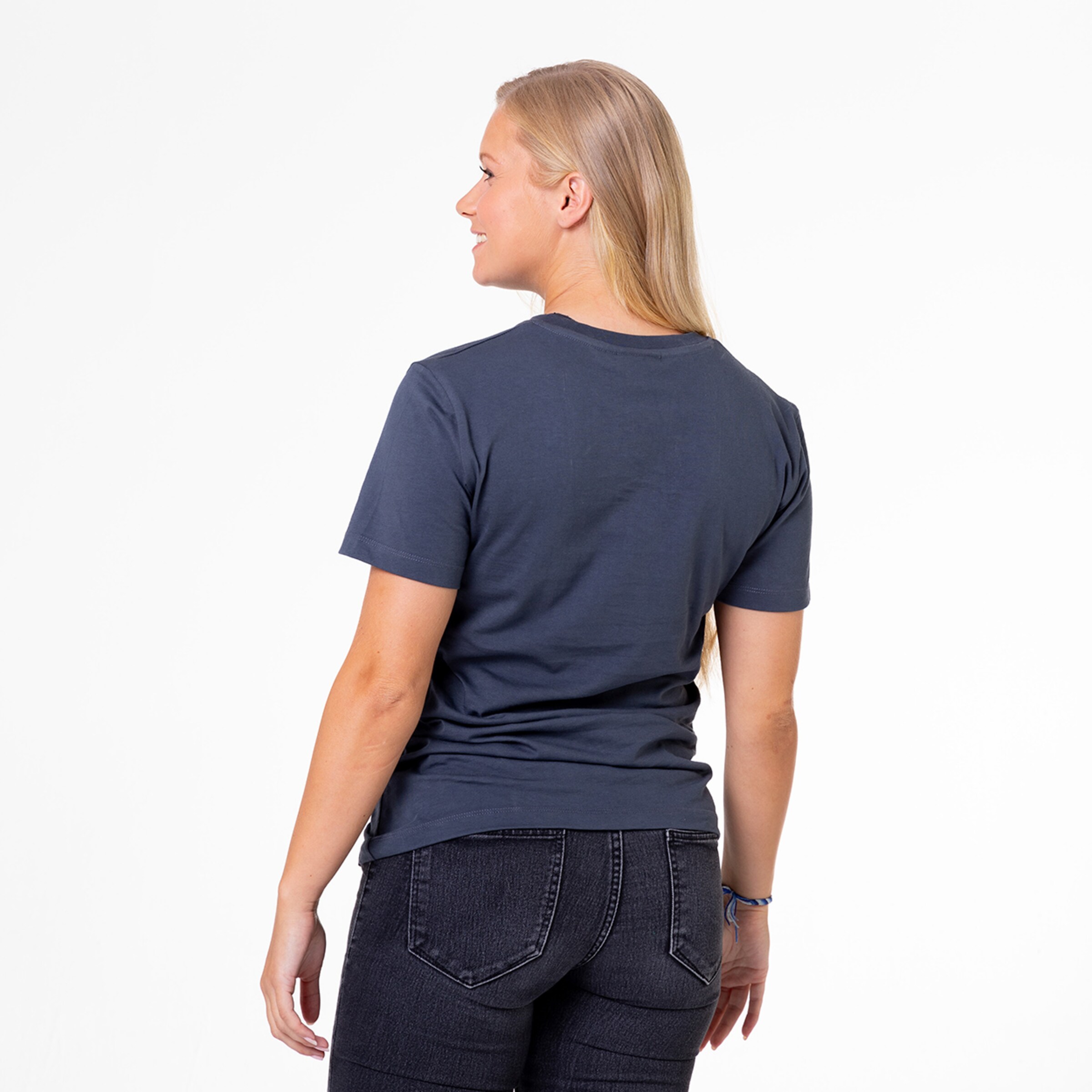 Eine Frau mit langen blonden Haaren trägt ein einfaches T-Shirt und sieht über ihre rechte Schulter zurück.