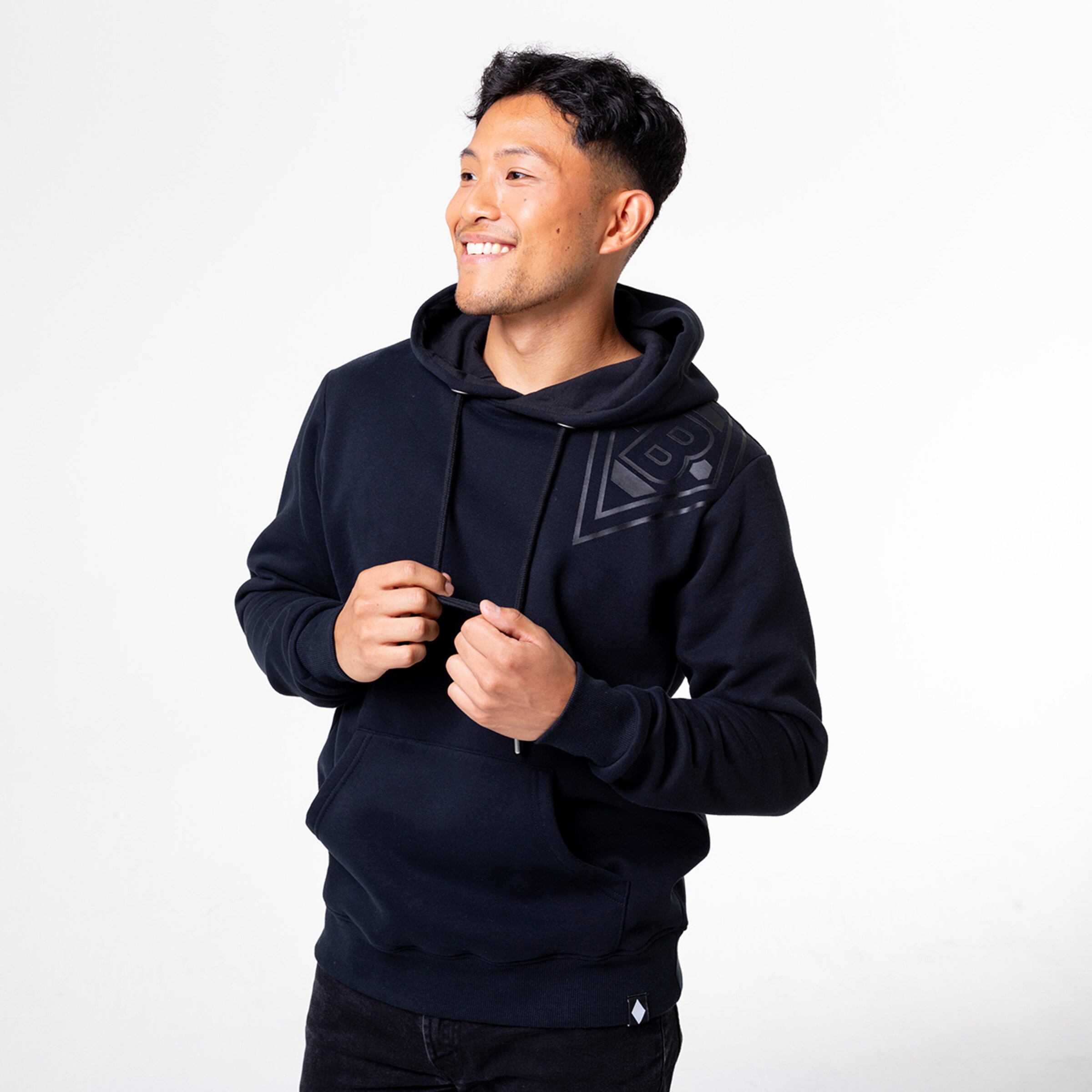Ein junger Mann trägt einen schwarzen Hoodie und lächelt während er seine Hände an der Vorderseite hält.