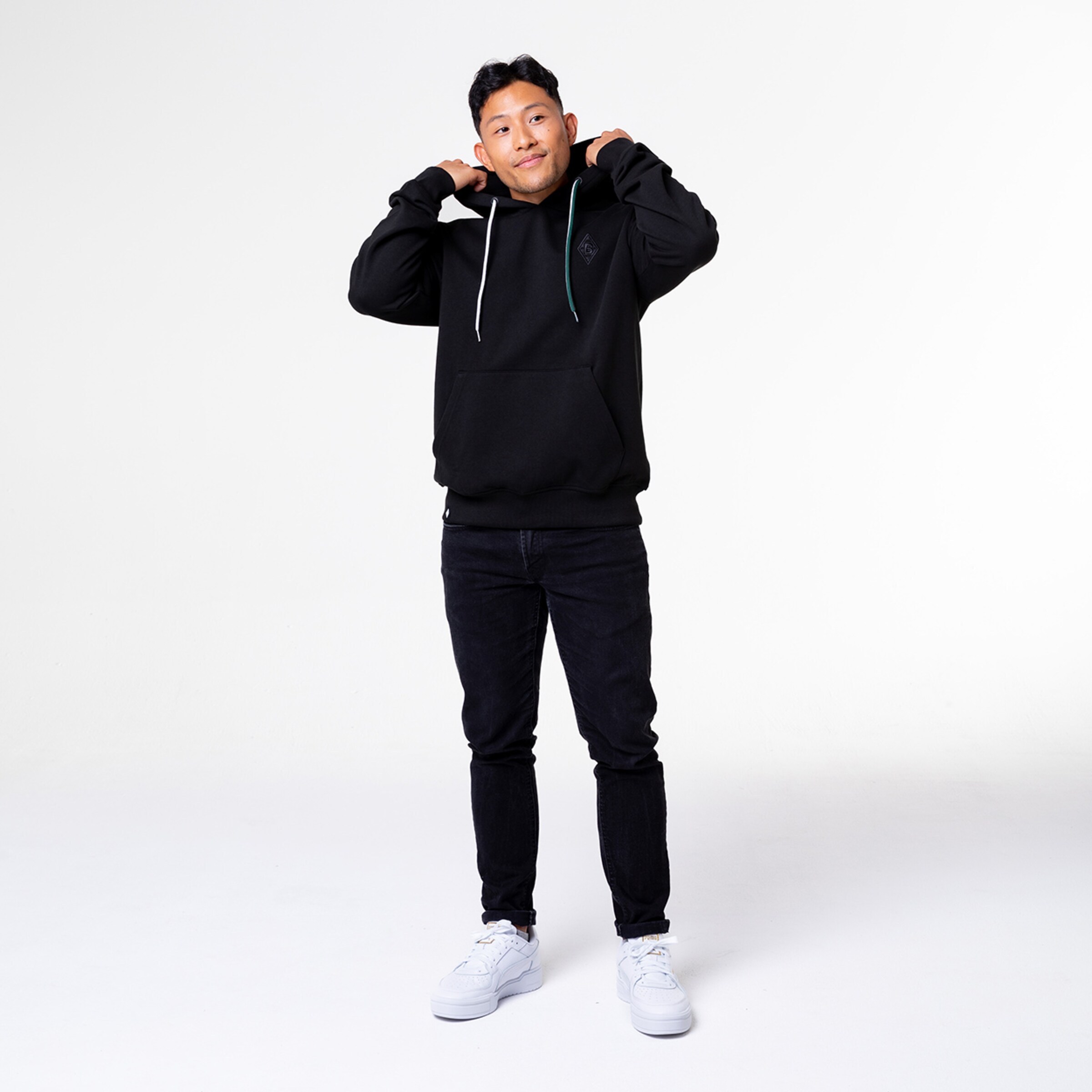 Ein Mann steht mit einem schwarzen Hoodie und schwarzen Jeans. Er lächelt und hält seinen Kapuzenpulli.