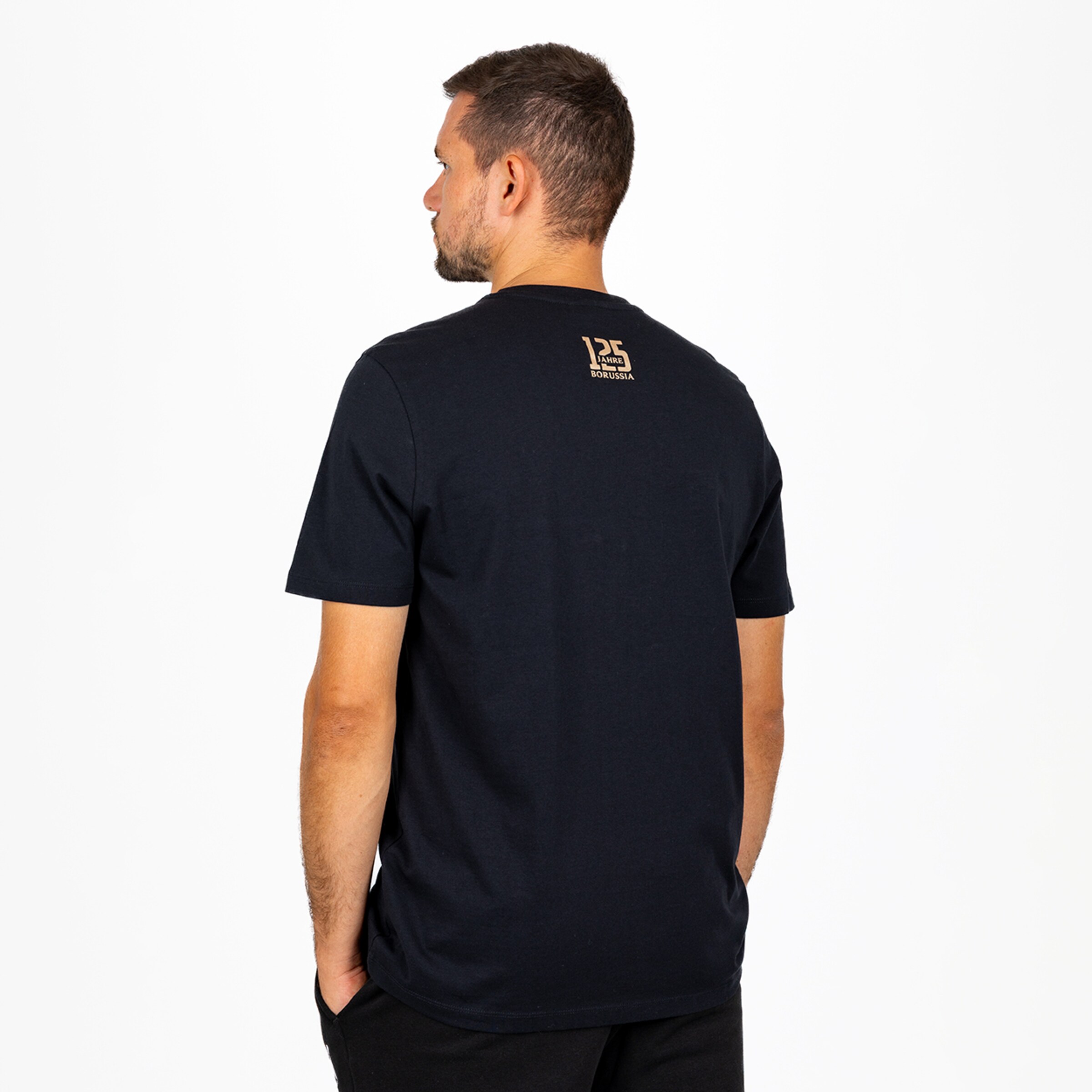 Rückansicht eines Mannes in schwarzem T-Shirt mit goldenem „125 Jahre Borussia“-Aufdruck im Nacken vor weißem Hintergrund.