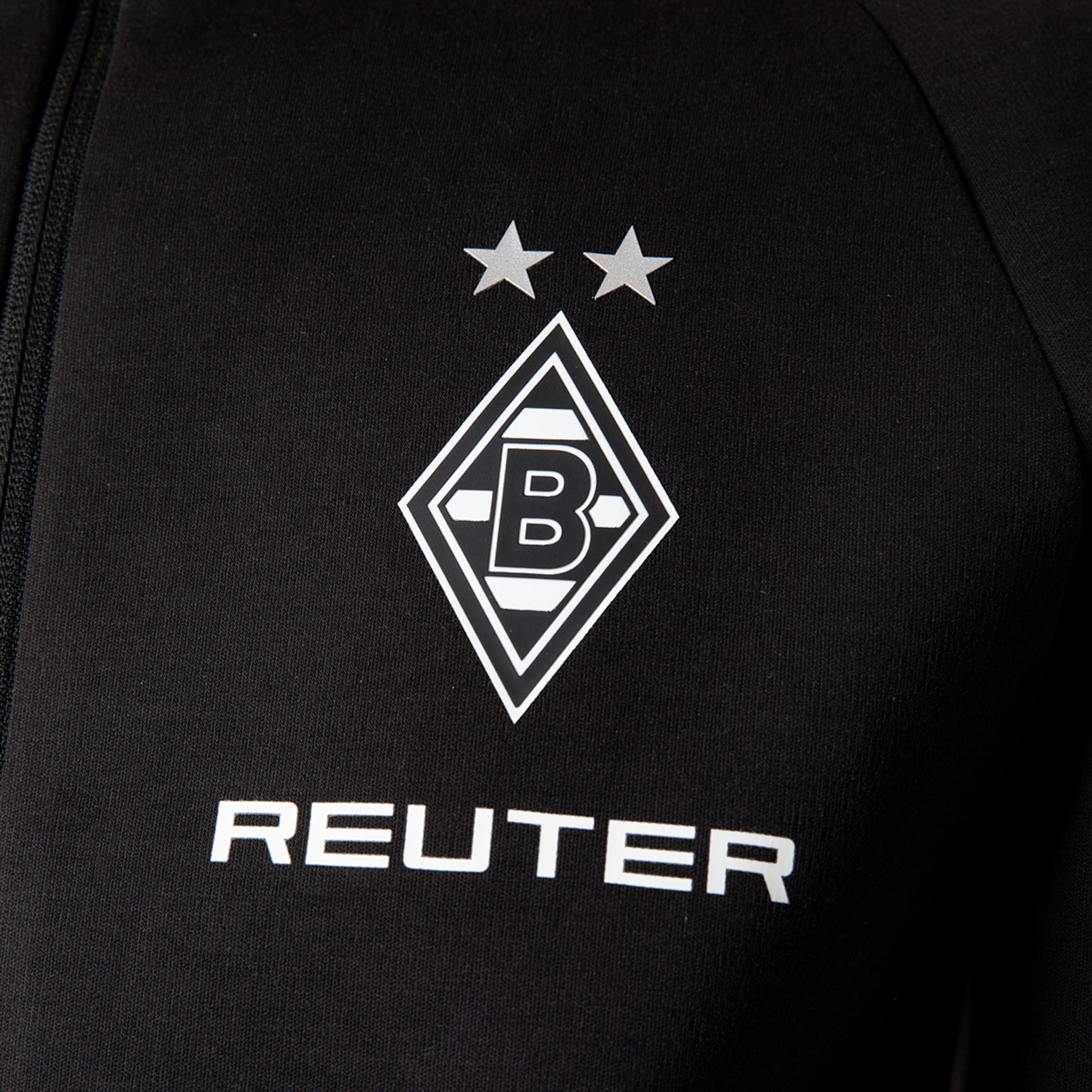 Nahaufnahme eines schwarzen Sportbekleidungsstücks, das das Logo von Borussia Mönchengladbach zeigt: eine Raute mit einem 'B' darin und zwei Sternen darüber. Darunter ist der Sponsor 'REUTER' in weißen Großbuchstaben zu sehen.