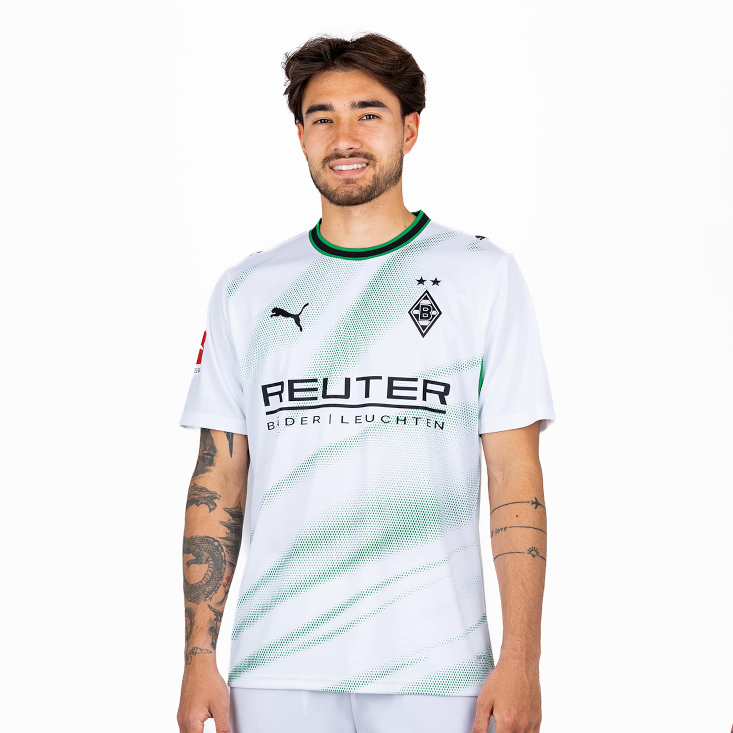 Ein lächelnder Mann im weißen Borussia Mönchengladbach Heimtrikot mit grünem Grafikmuster und Tattoos vor einem neutralen weißen Hintergrund.
