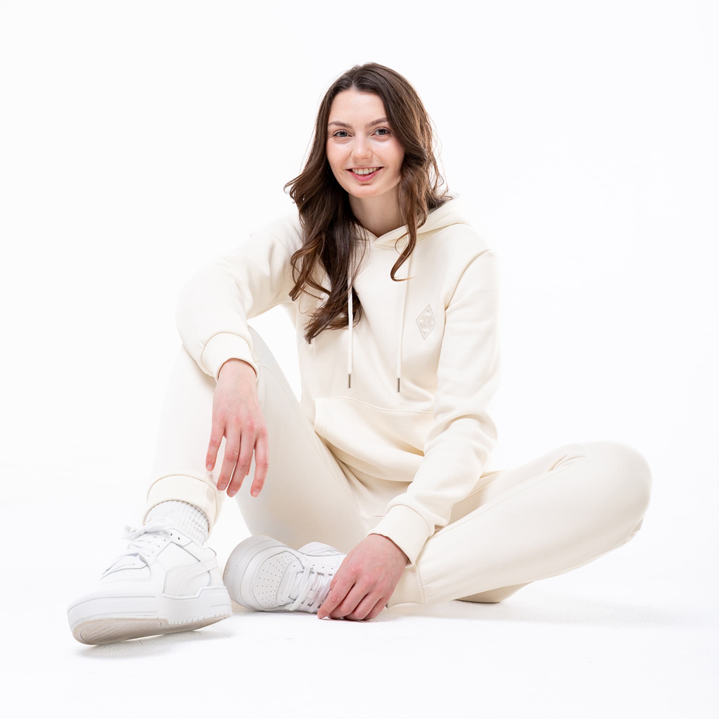 Eine Frau sitzt entspannt in einem weißen Trainingsanzug mit Kapuze und Sneakers.