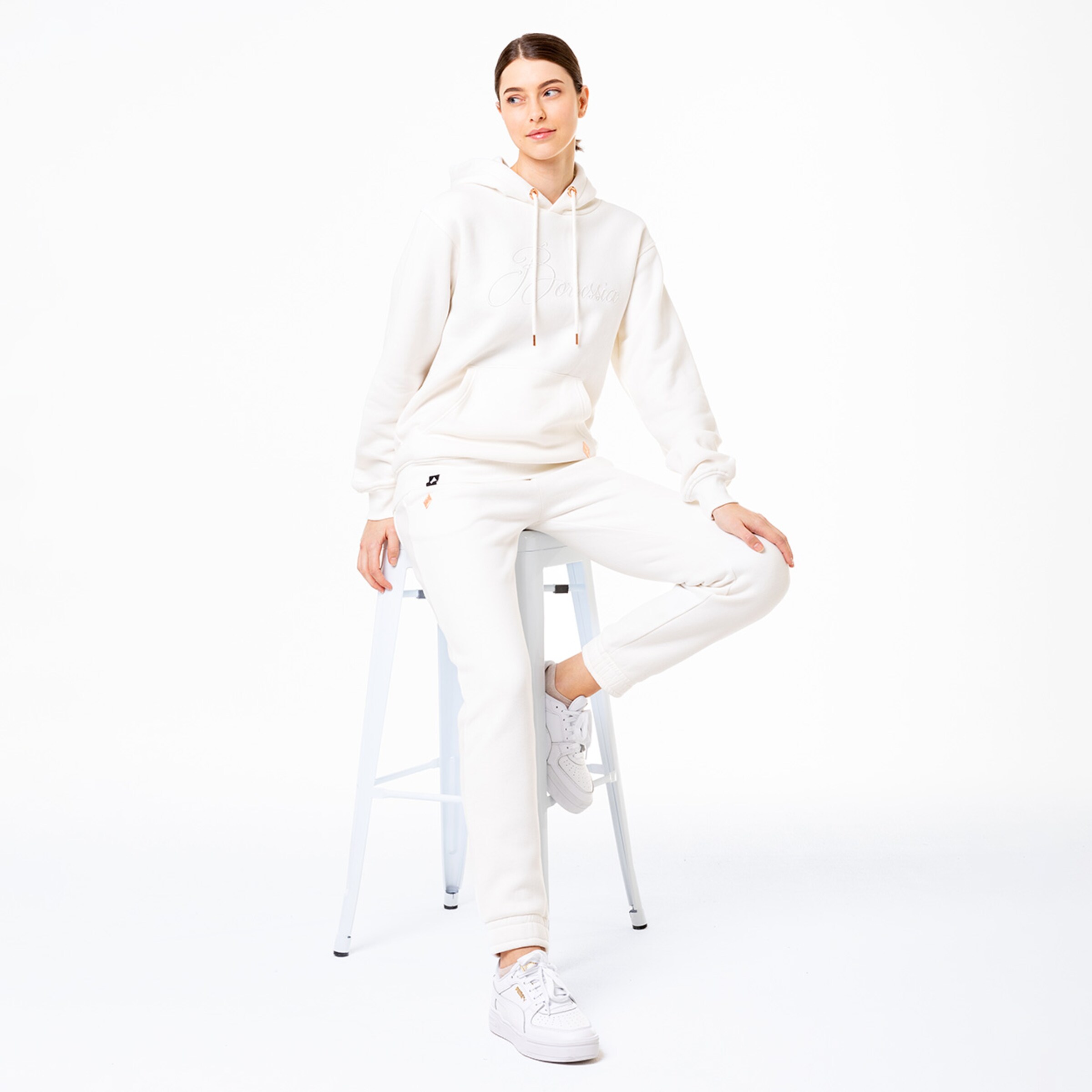 Eine Frau sitzt auf einem Hocker und trägt einen weißen Hoodie und passende Jogginghose.