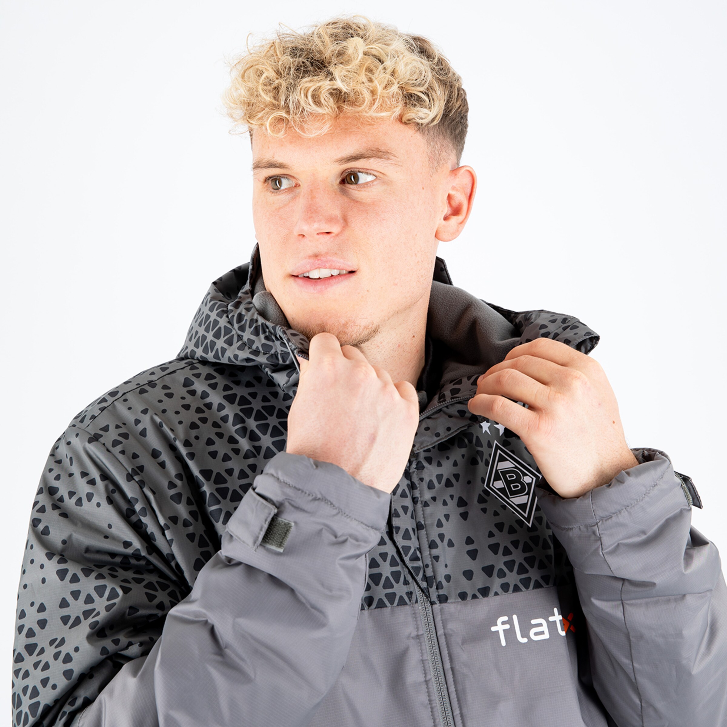 Ein junger Mann mit lockigem Blondhaar trägt eine graue Jacke mit einem Muster und zieht den Reißverschluss bis zum Kinn hoch.