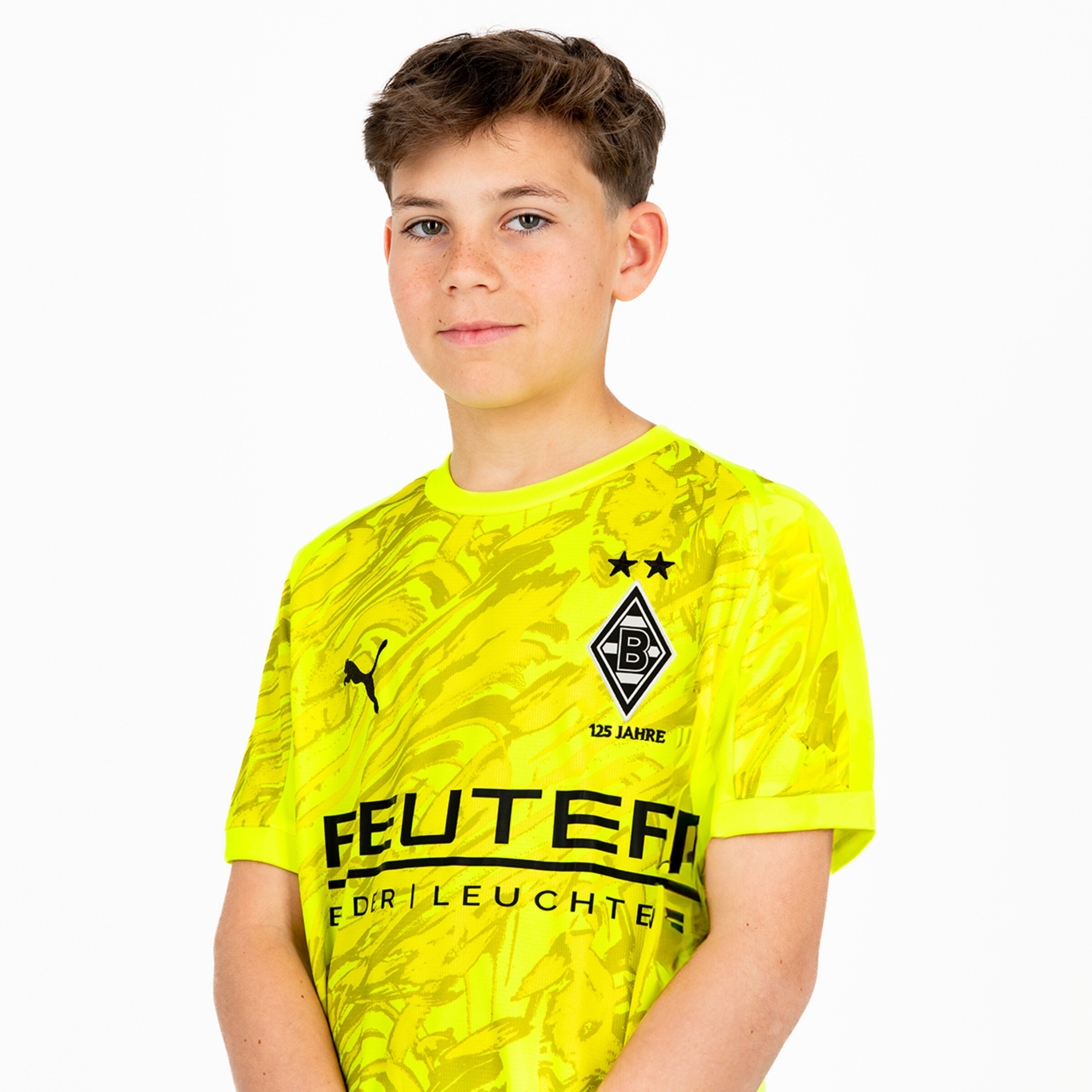 Ein Junge steht in einem leuchtend gelben Borussia Mönchengladbach Trikot und lächelt leicht.