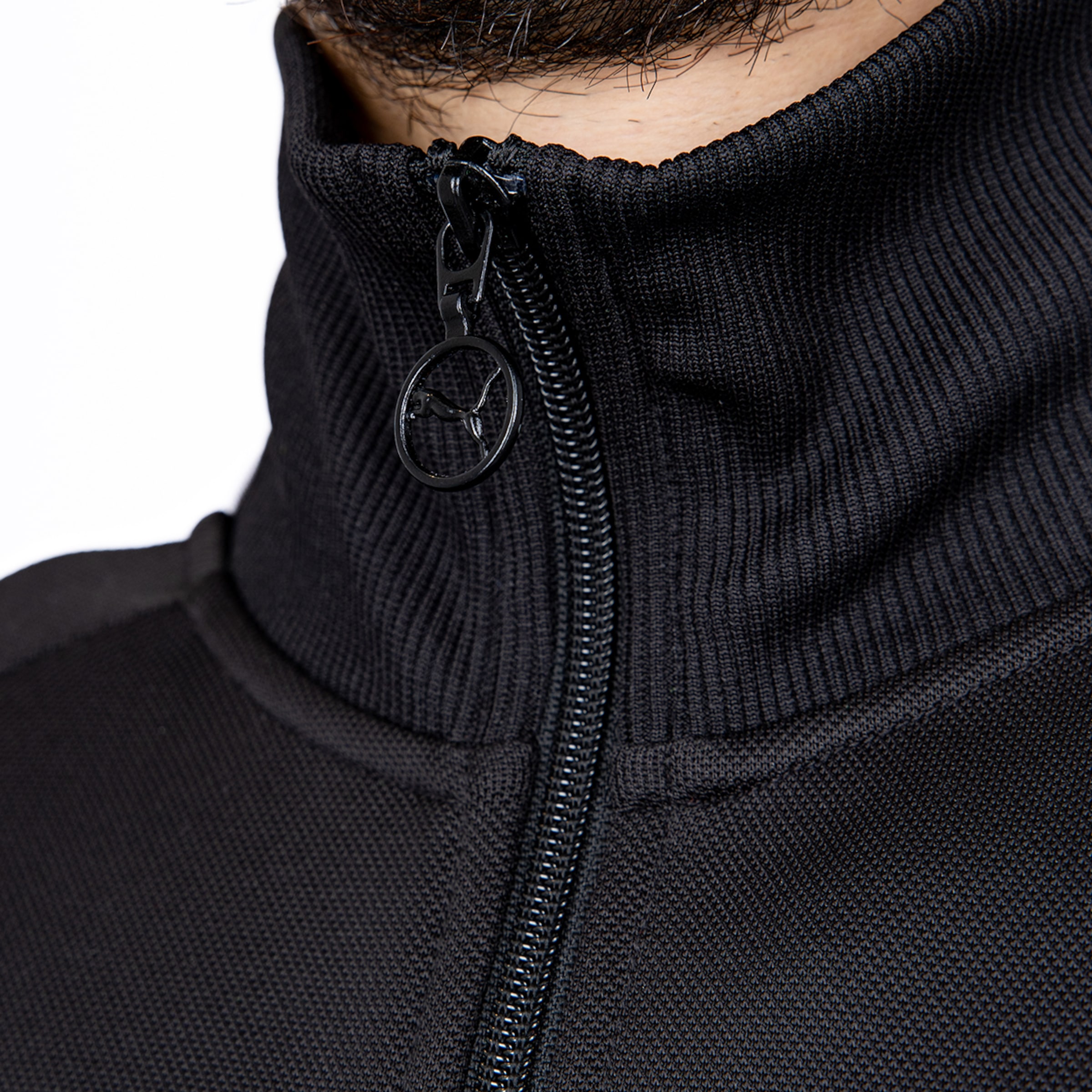 Nahaufnahme eines schwarzen Stehkragens einer Jacke mit Reißverschluss und rundem Puma-Logo-Anhänger.