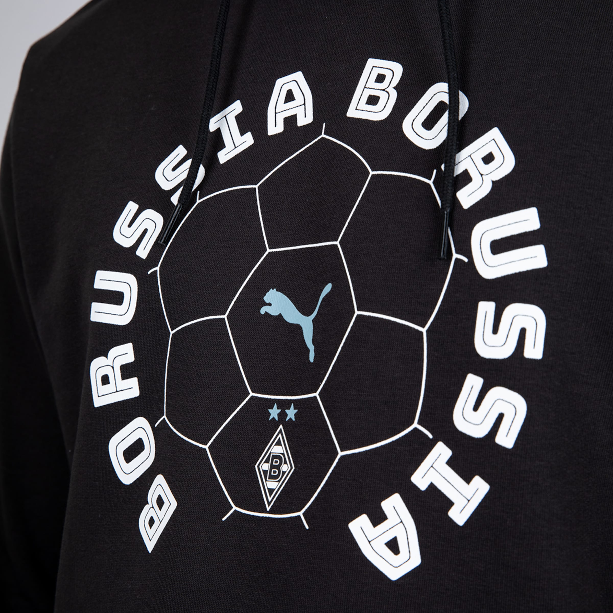 Nahaufnahme eines schwarzen Hoodies mit weißem Rund-Aufdruck: „BORUSSIA“-Schriftzug, Fußballmuster, hellblaues Puma-Logo, zwei Sterne und das Borussia Mönchengladbach Wappen.