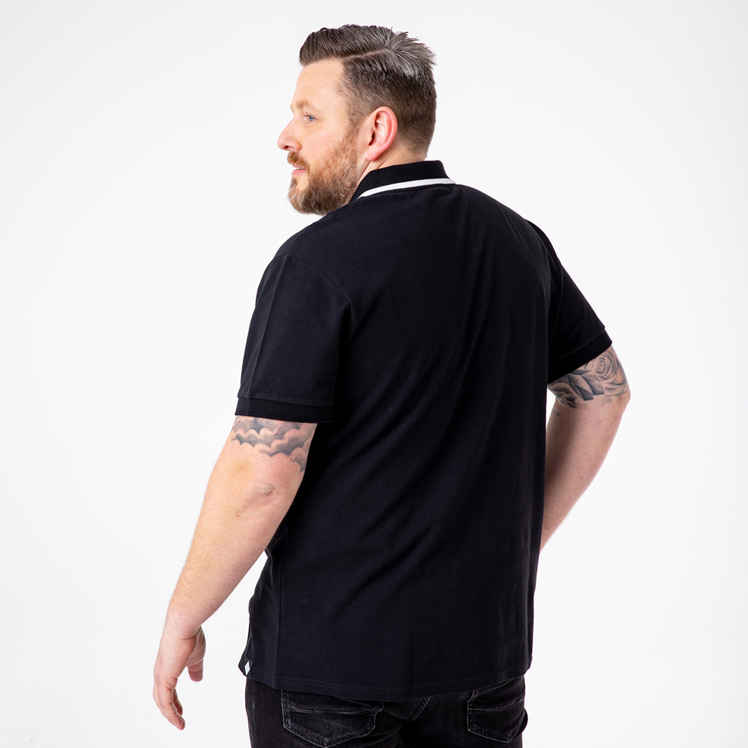 A man with short hair and tattoos is wearing a black polo shirt and looking over his left shoulder.