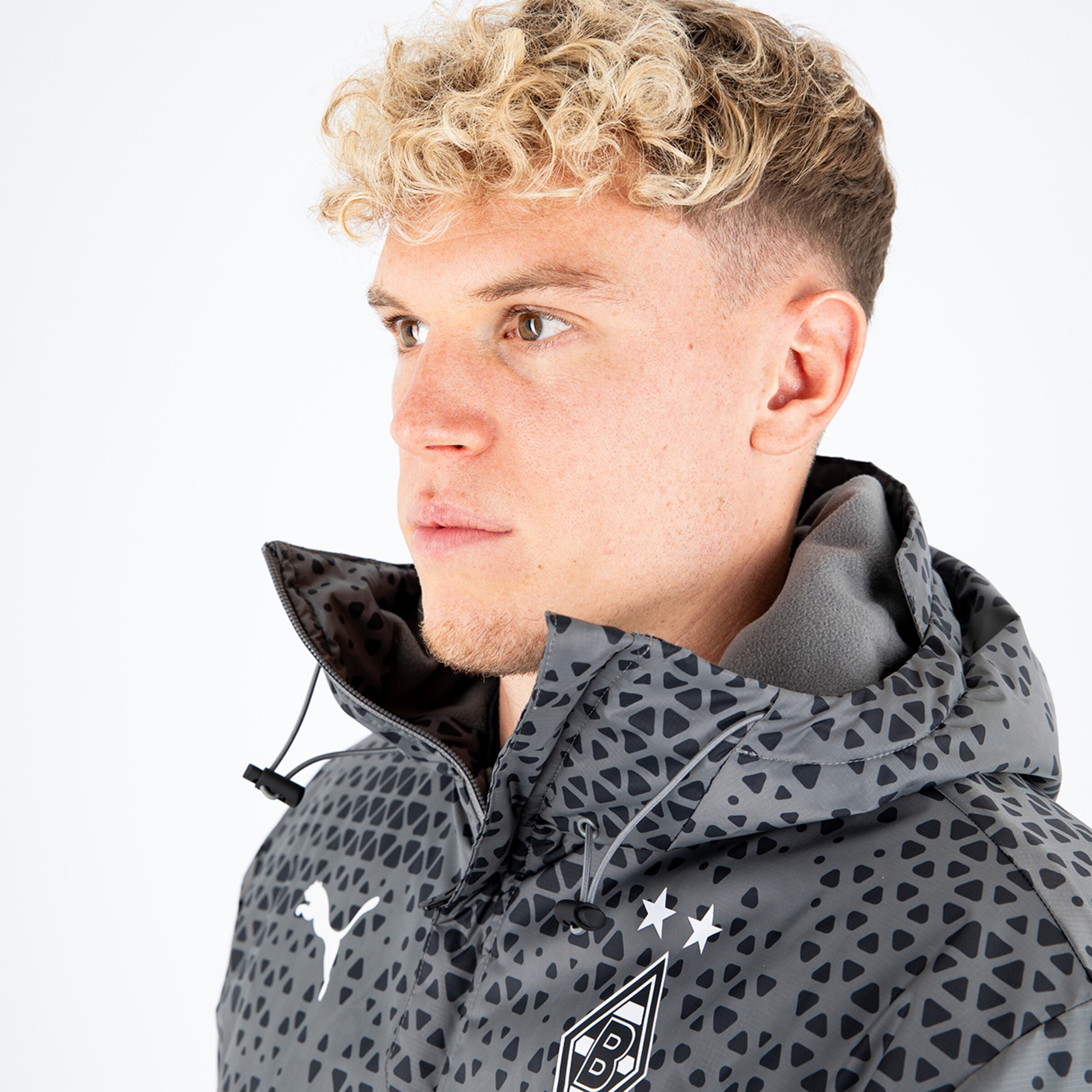 Mann mit lockigen blondem Haar trägt eine sportive graue Jacke mit Puma-Logo und Borussia Mönchengladbach-Emblem.
