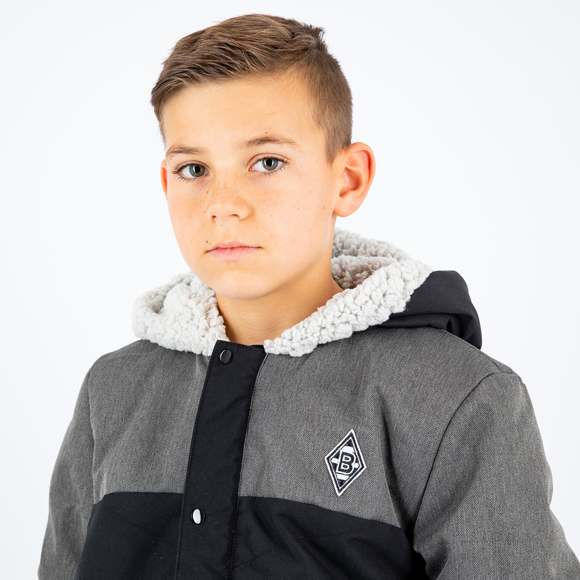 Boy with short hair in gray jacket looking forward logo on the jacket visible.