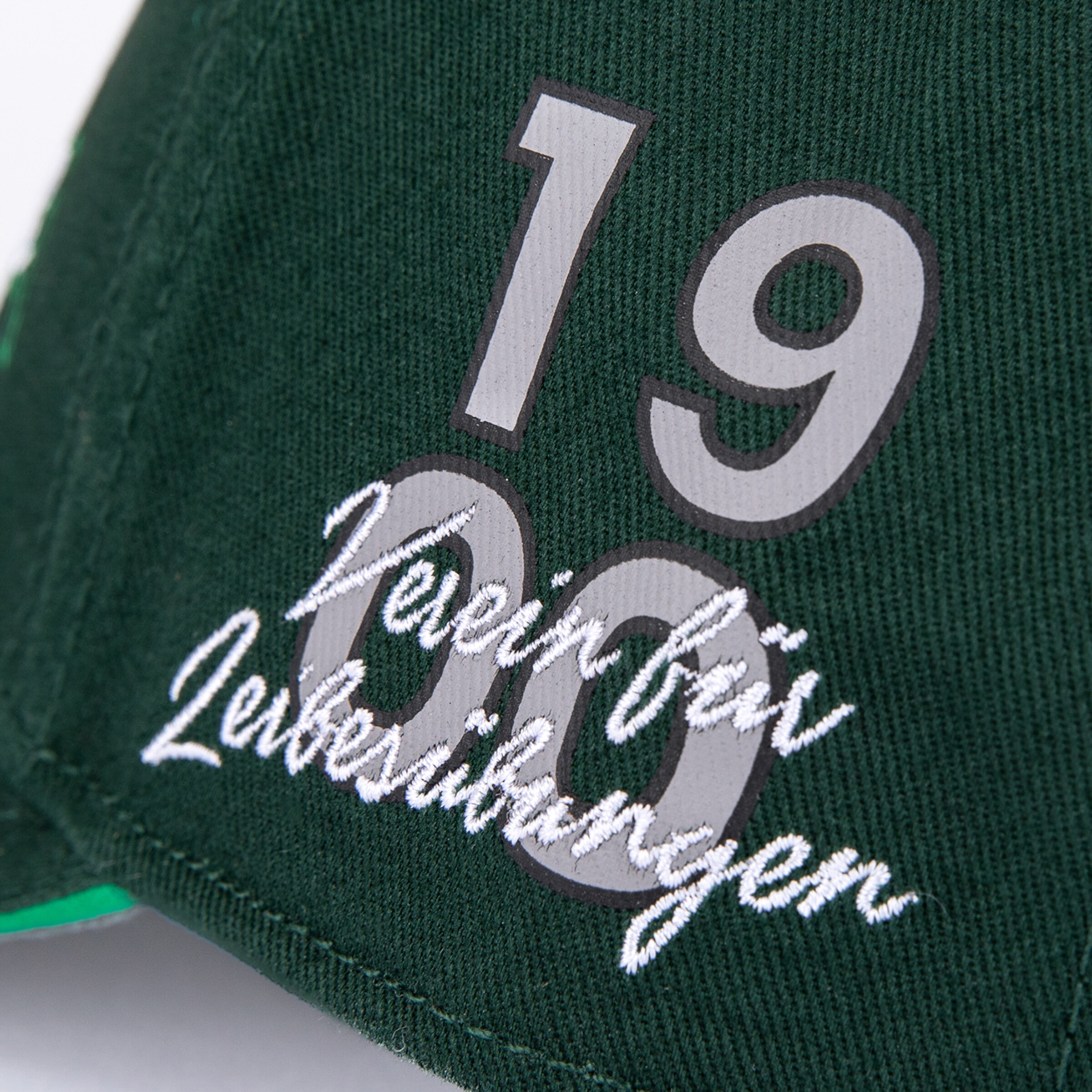 Close-up of a green cap with silver number 1900 and white embroidered cursive text reading 'Verein für Leibesübungen'.