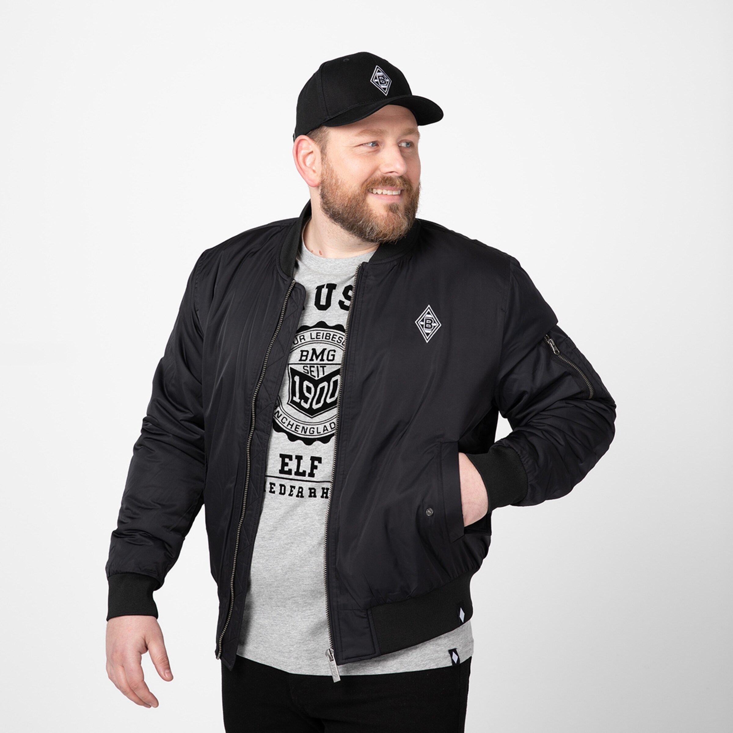 Man in a black bomber jacket and black cap against a neutral background.