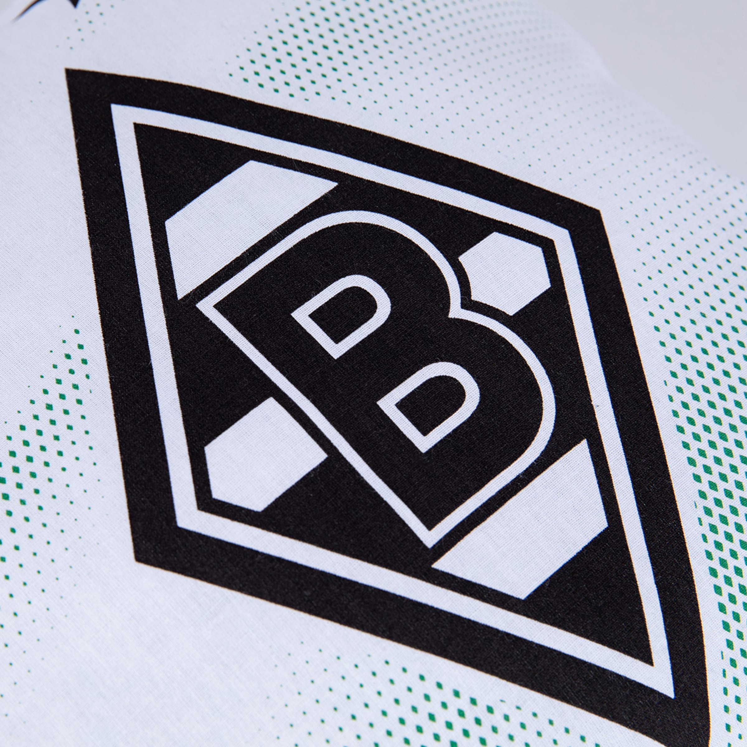 Nahaufnahme des Borussia Mönchengladbach Logos: Schwarze Raute mit weißem „B“ auf weißem Stoff mit grünem Punktmuster.