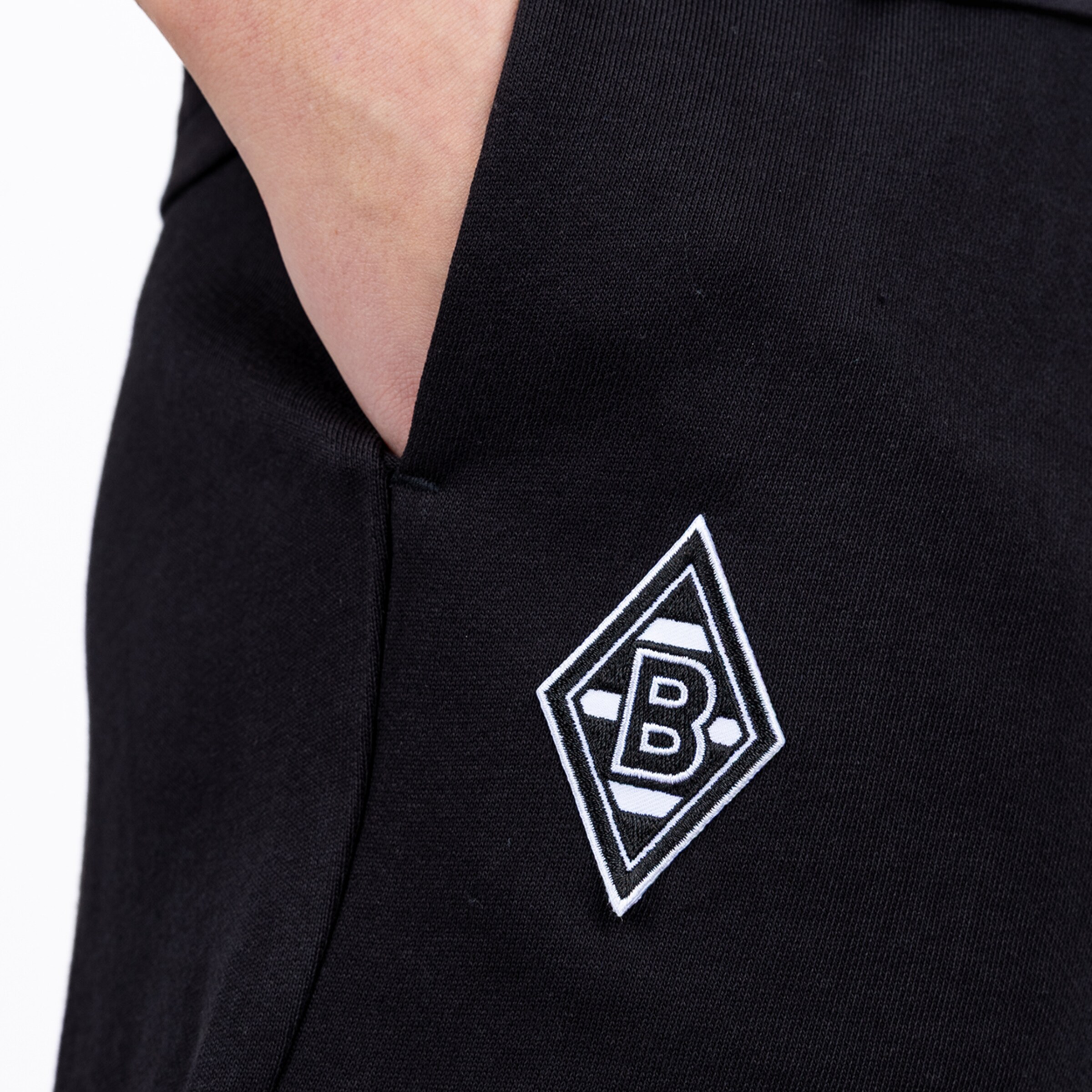 Nahaufnahme einer schwarzen Jogginghose mit Hand in der Tasche. Unter der Seitentasche prangt das gestickte Logo von Borussia Mönchengladbach.