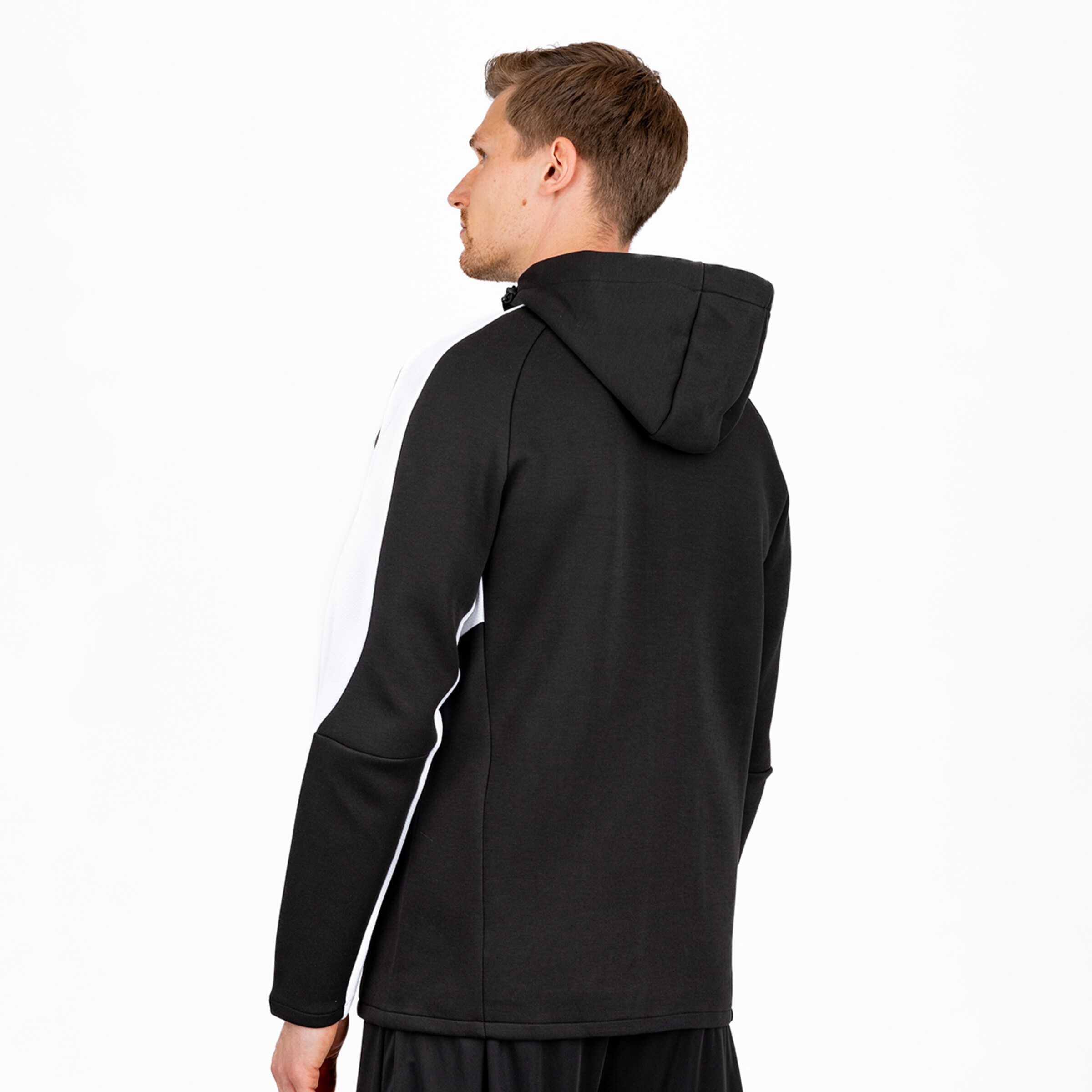 Ein Mann, von hinten gesehen und leicht nach links gedreht, trägt eine schwarze Kapuzenjacke mit weißen Streifen an den Ärmeln. Er hat kurzes braunes Haar und einen leichten Bart. Der Hintergrund ist weiß.