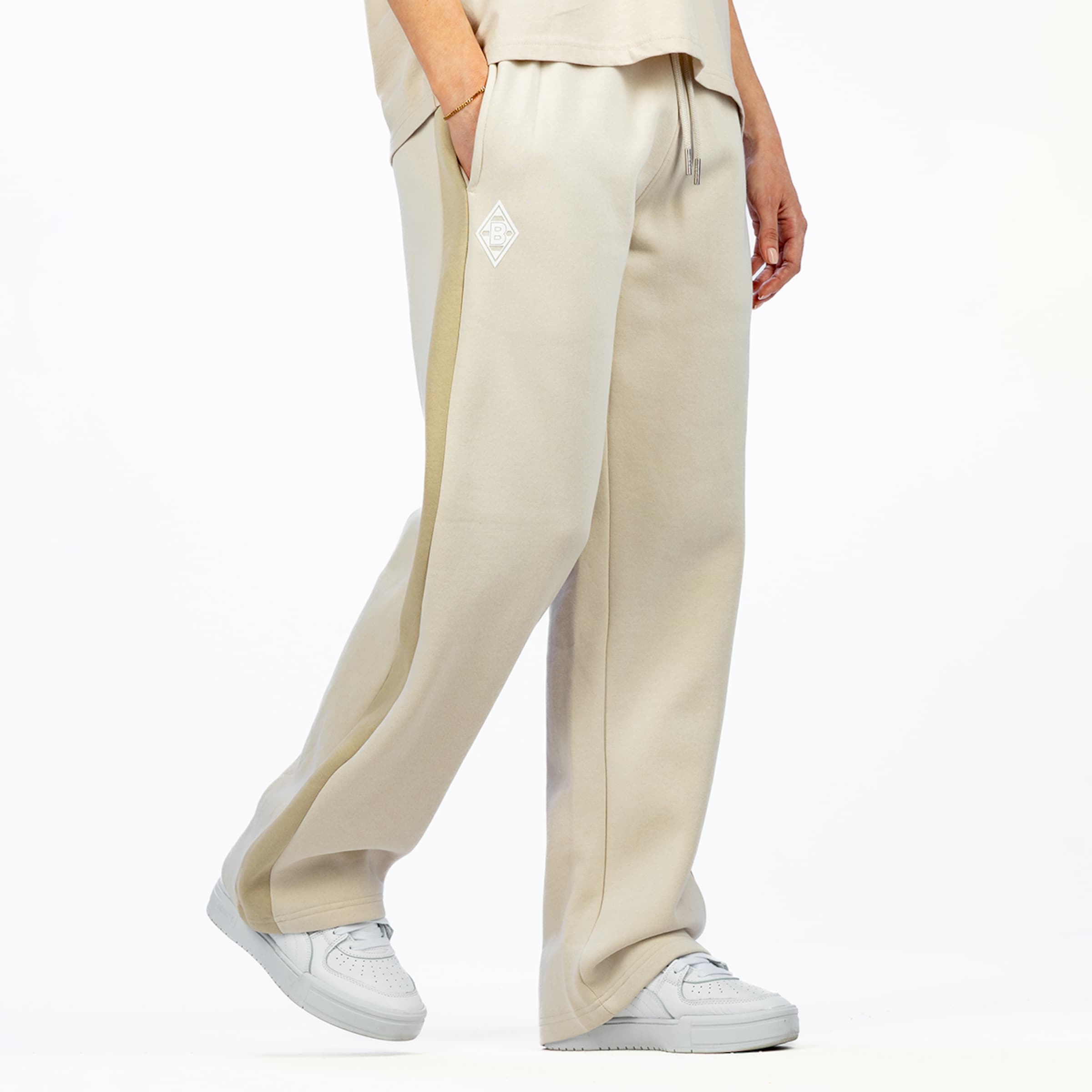 A person wears beige sweatpants with a light side stripe, white sneakers, and a gold bracelet. A white logo is visible on the left thigh.