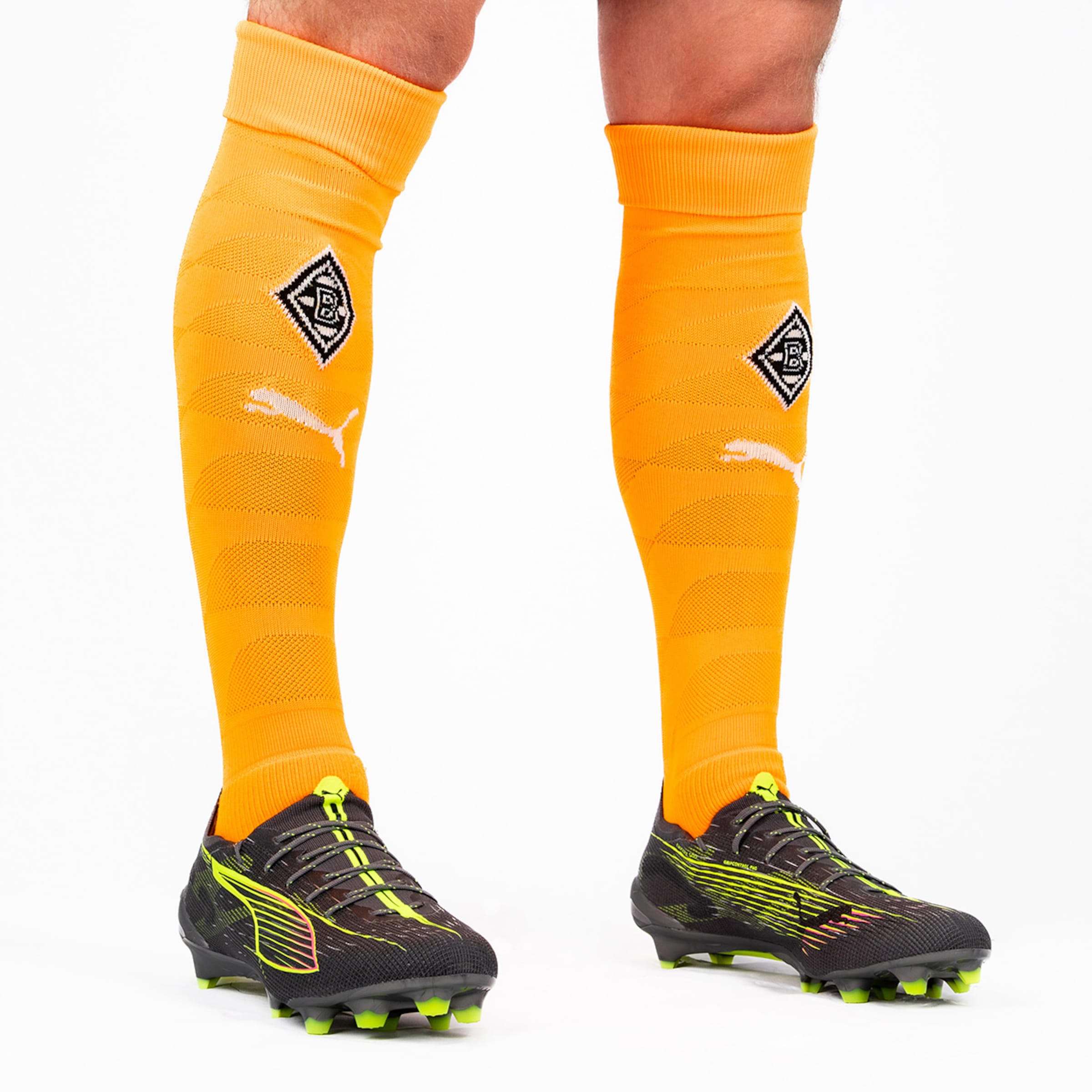 Ein Spieler trägt orangefarbene Schienbeinschoner mit dem Borussia Mönchengladbach Logo und schwarze Fußballschuhe.