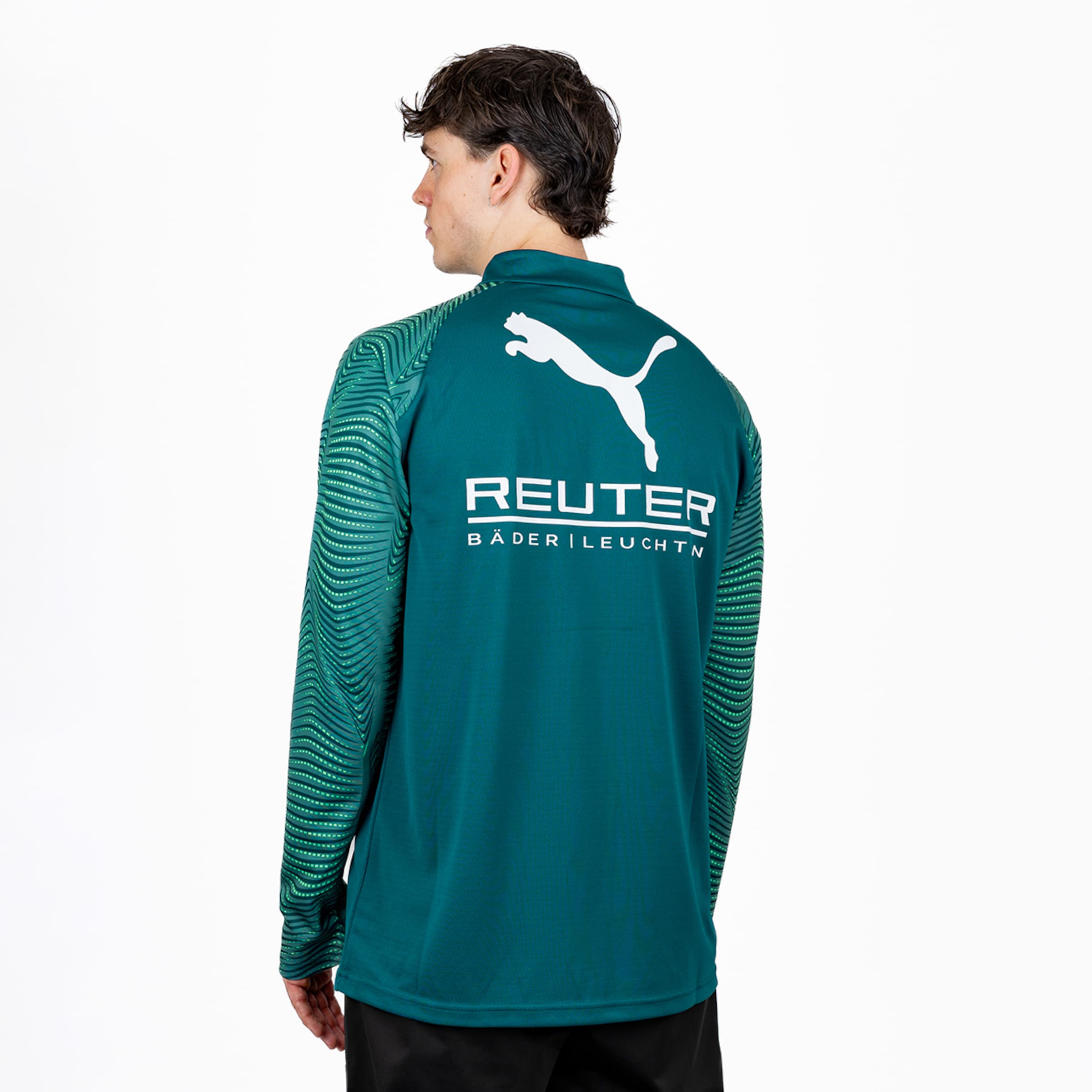 Mann von hinten, nach links blickend, in dunkelgrünem Langarmshirt. Auf dem Rücken sind ein weißes Puma-Logo und der Schriftzug „REUTER BÄDER | LEUCHTEN“ zu sehen. Die Ärmel haben ein gewelltes Streifenmuster.