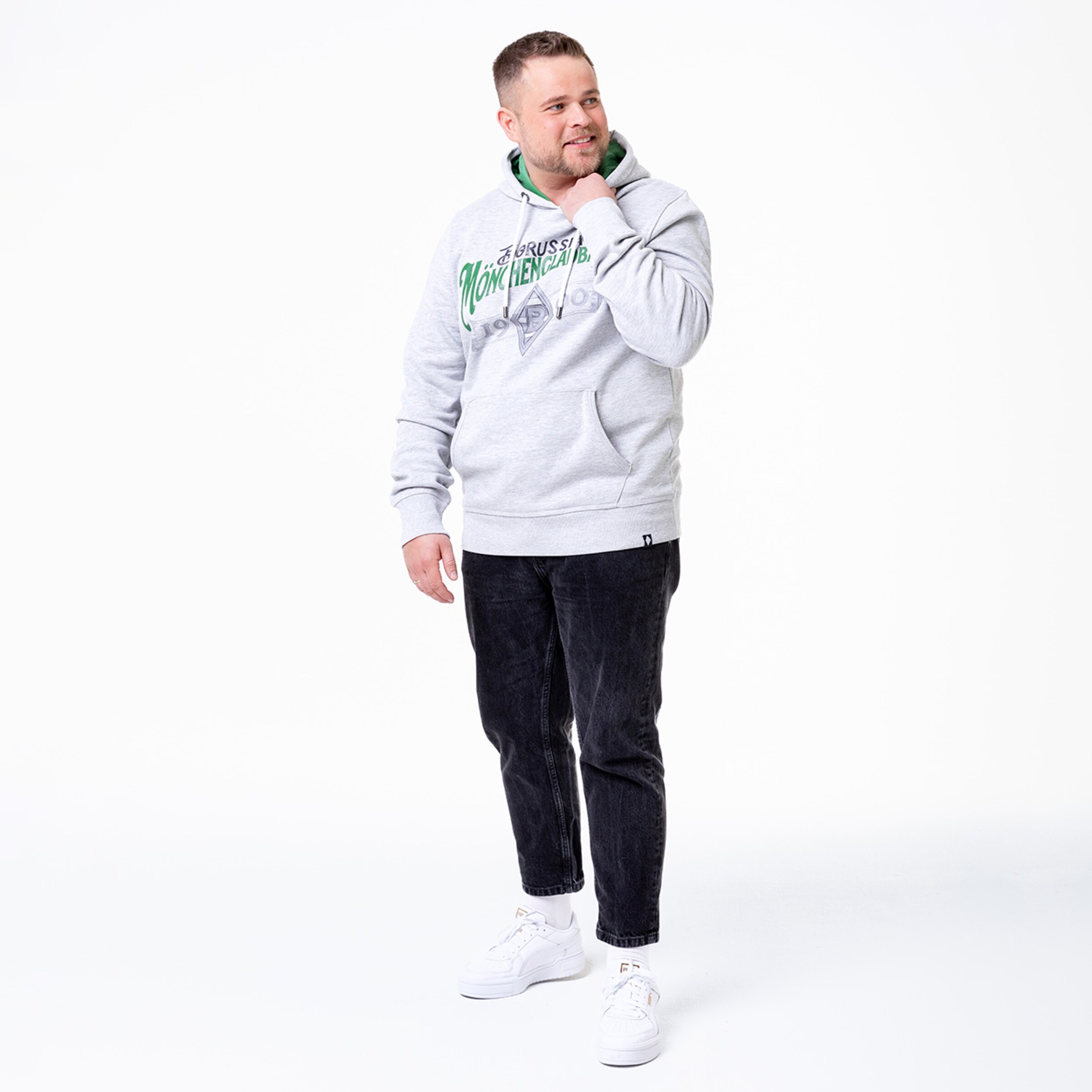 Mann in grauem Hoodie mit Borussia Mönchengladbach-Logo und schwarzen Jeans lächelt.