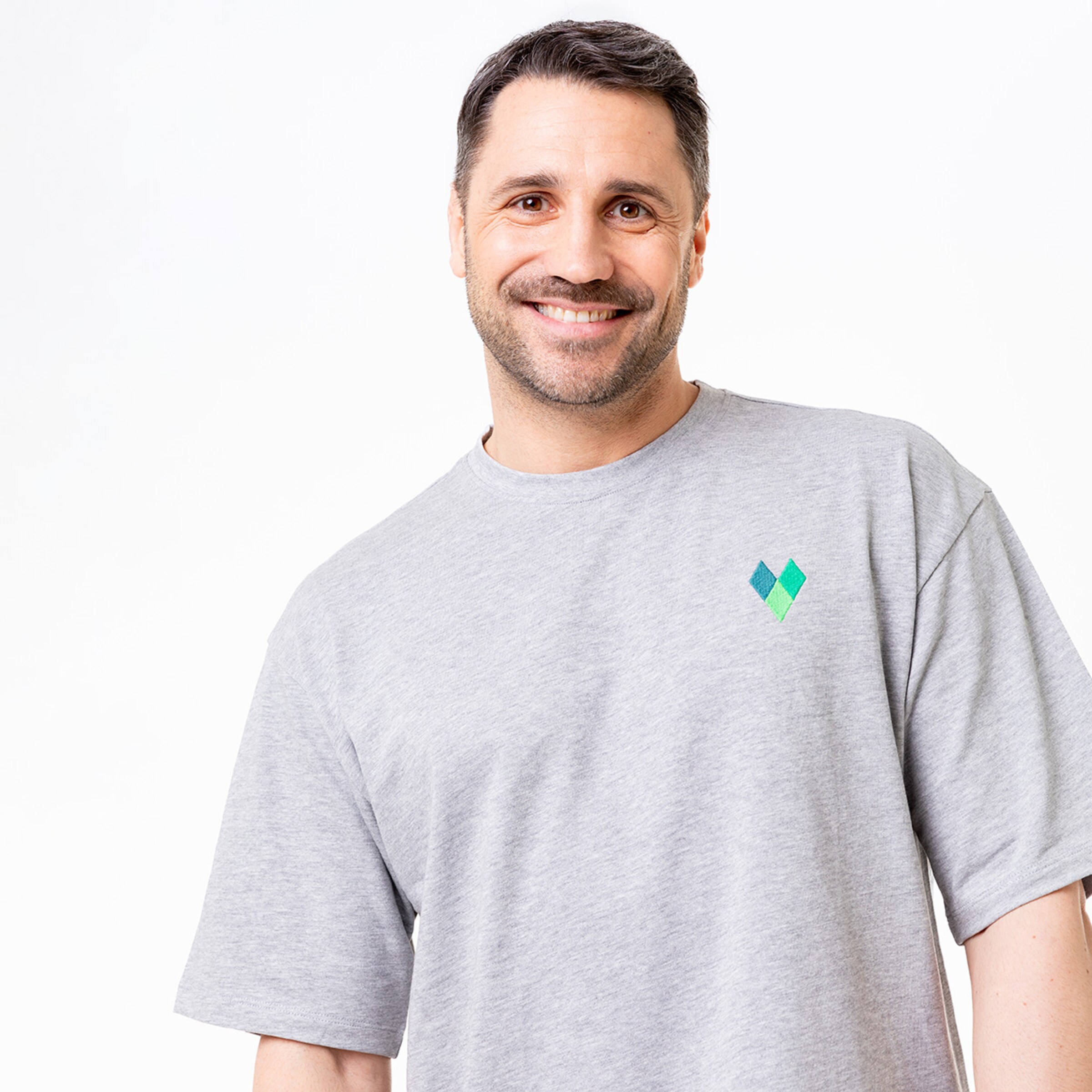 Ein Mann lächelt trägt ein graues T-Shirt mit einem kleinen grünen Logo auf der Brust.