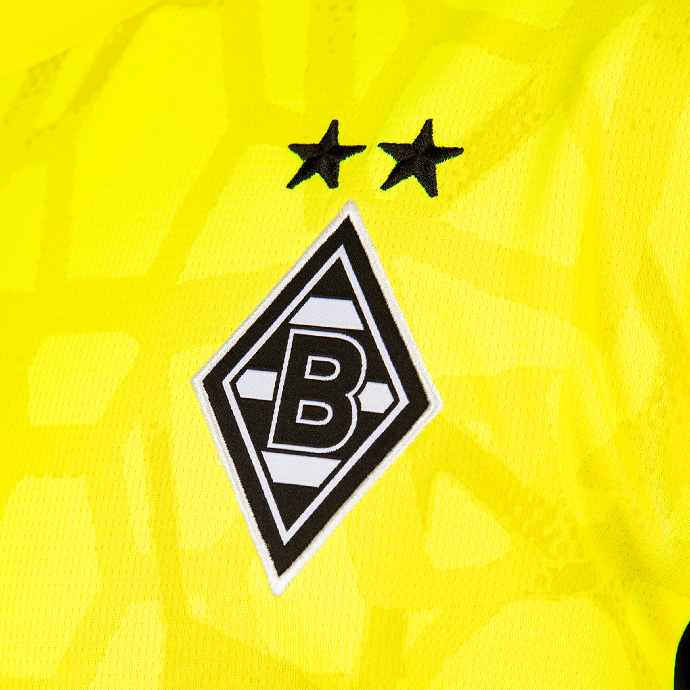 Logo von Borussia Mönchengladbach auf gelbem Trikot: Schwarz-weiße Raute mit dem Buchstaben B und zwei schwarzen Sternen darüber.