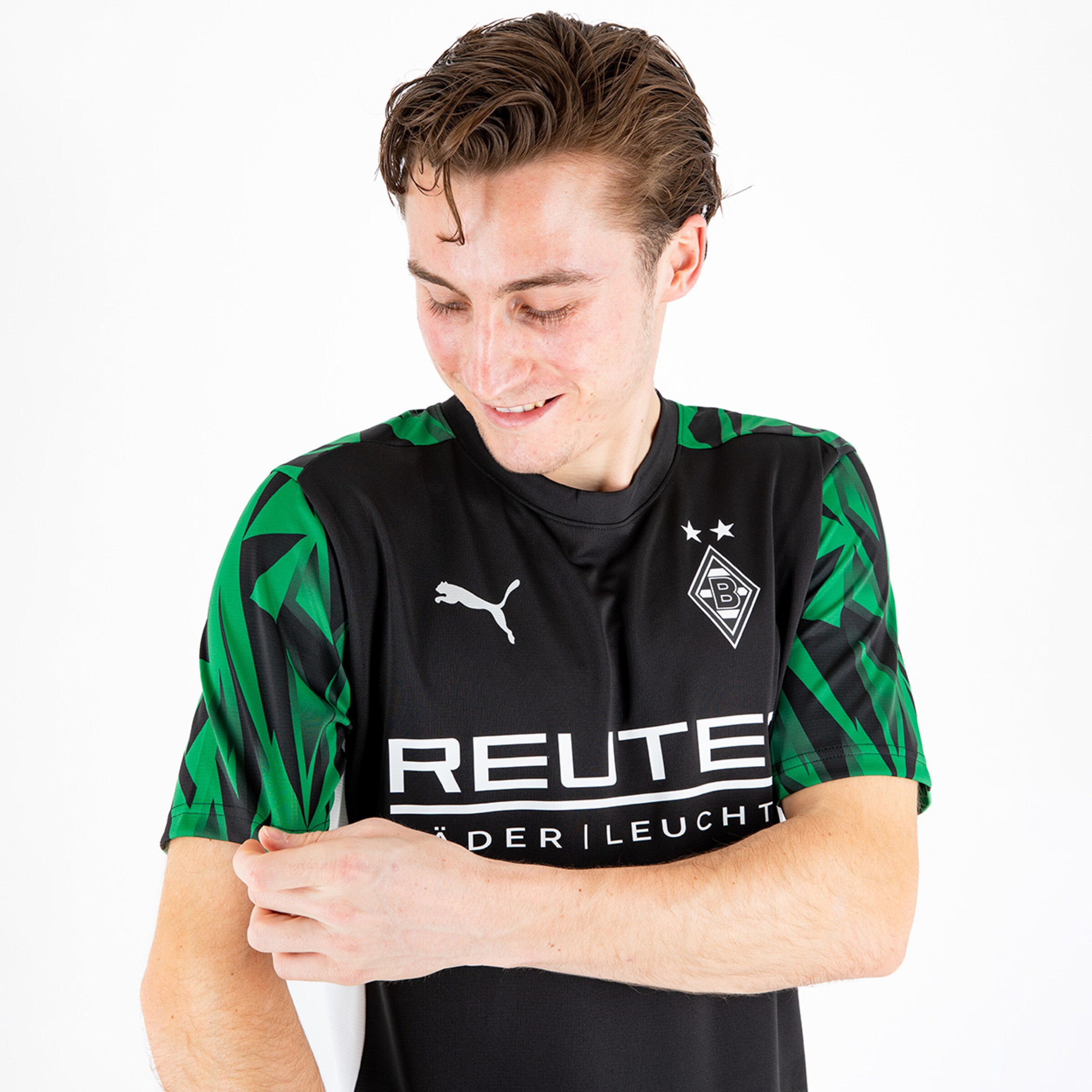 Ein junger Mann in einem Fußballtrikot mit grünem Muster zeigt auf seinen Ärmel.