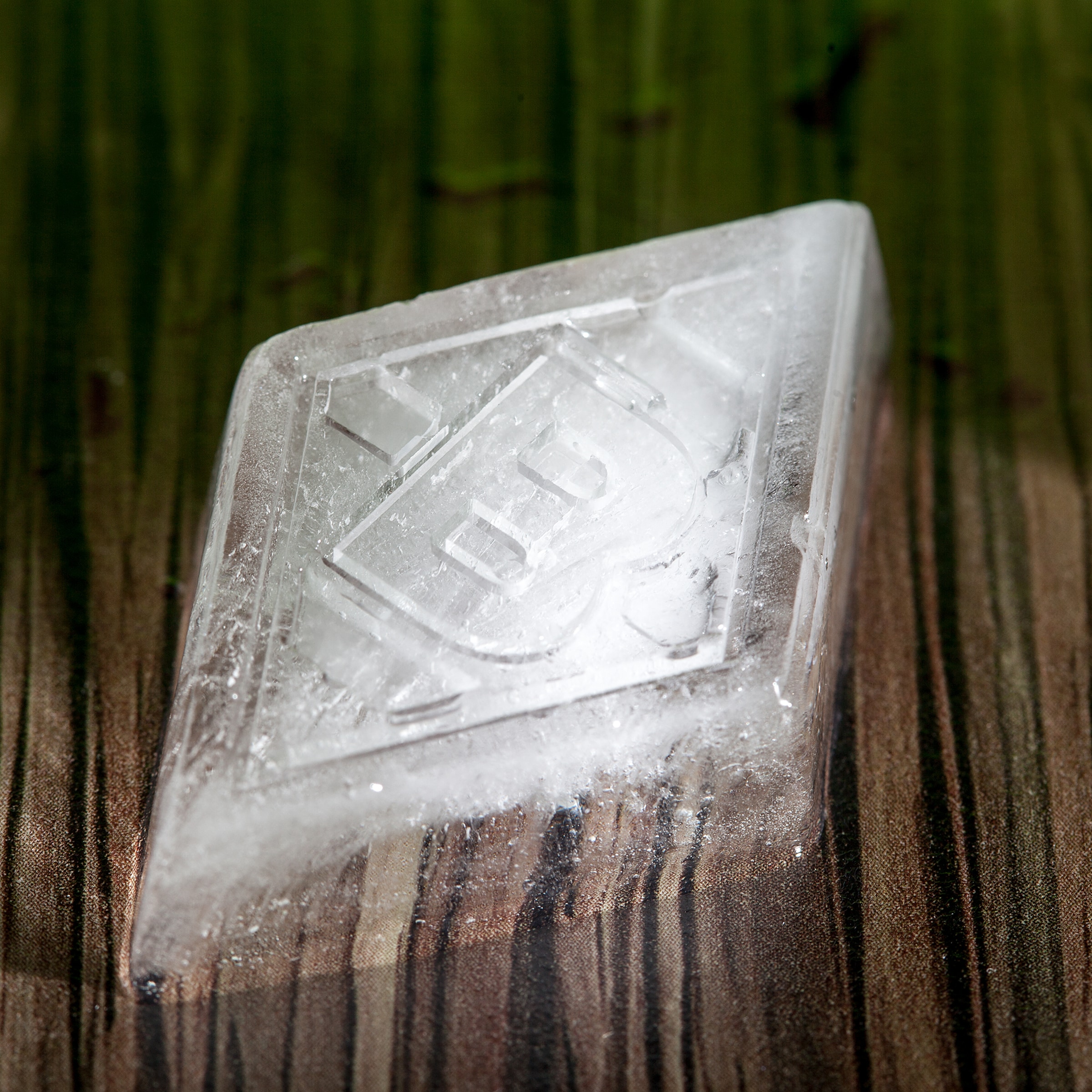 Rautenförmiger Eiswürfel mit eingeprägtem Logo auf einer dunklen Holzoberfläche. Das Eis ist mattiert und zeigt feine kristalline Strukturen.