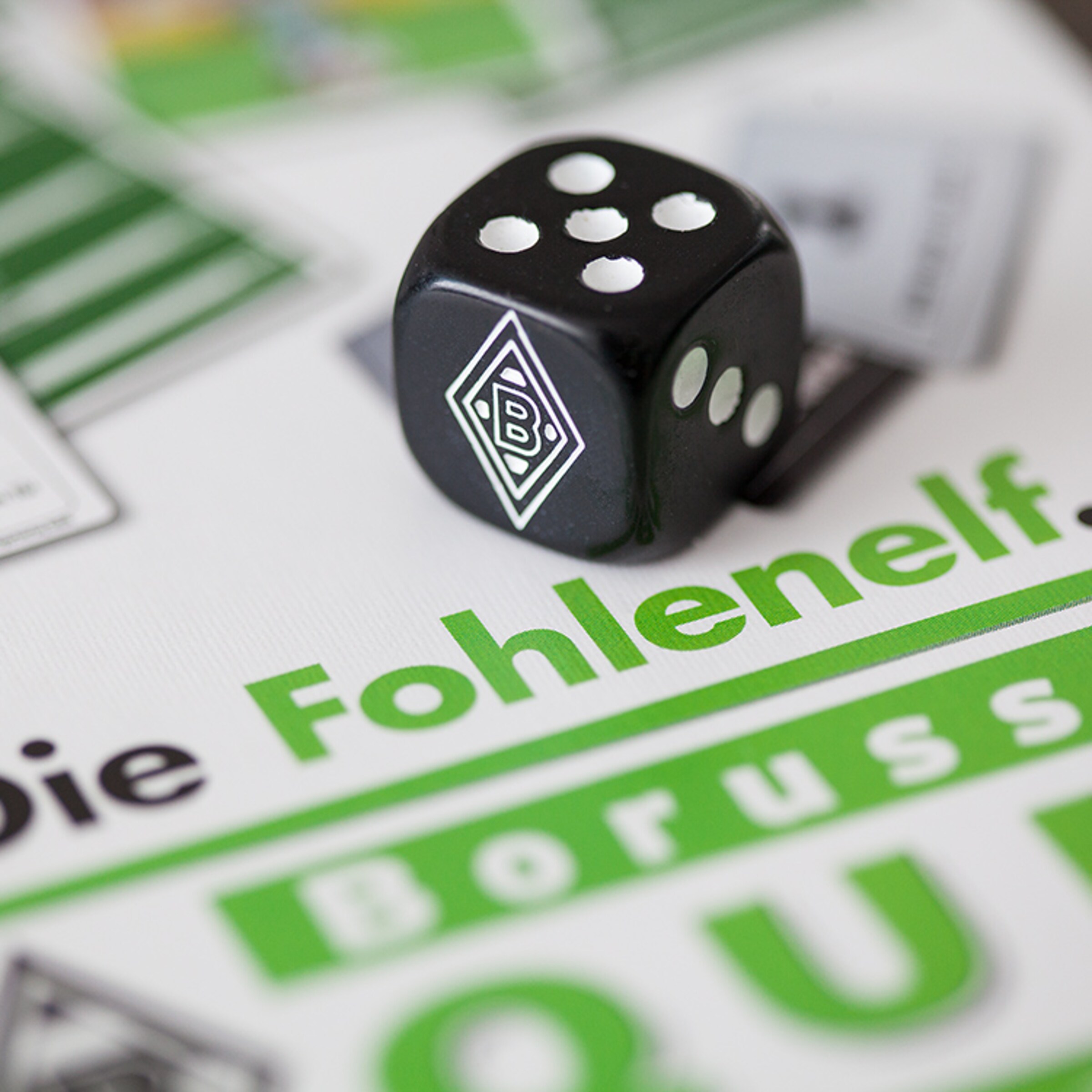 Schwarzer Würfel mit weißen Punkten und Borussia Mönchengladbach-Logo auf einem Spielbrett.