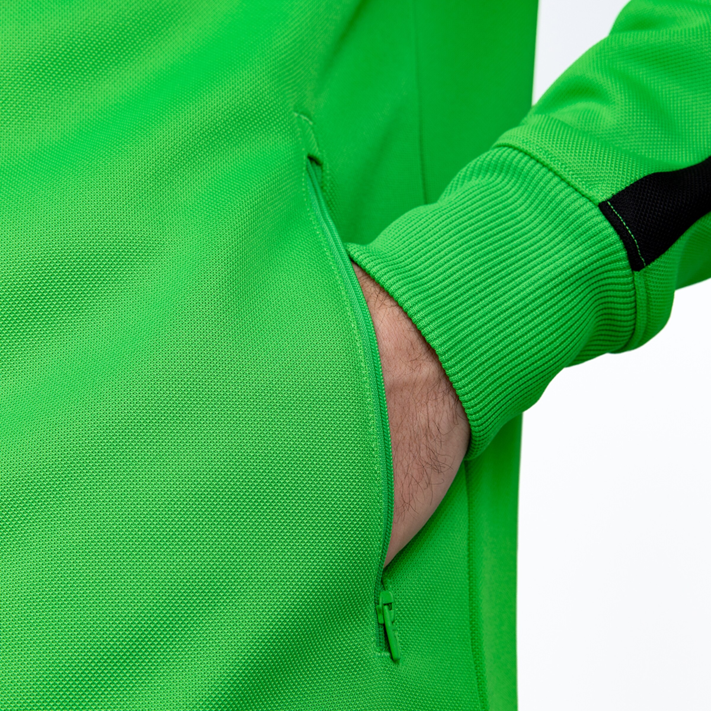 Nahaufnahme einer grünen Sportjacke mit Reißverschlusstasche und Rippbündchen am Ärmel. Eine Hand steckt in der Tasche.